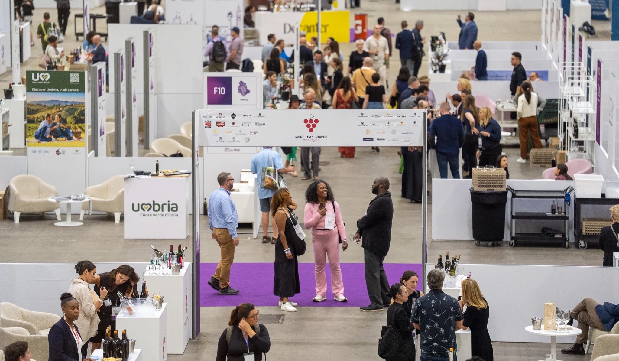 A crowded indoor event or exhibition with booths, tables, and people networking, with some people tasting wine at a wine tasting booth