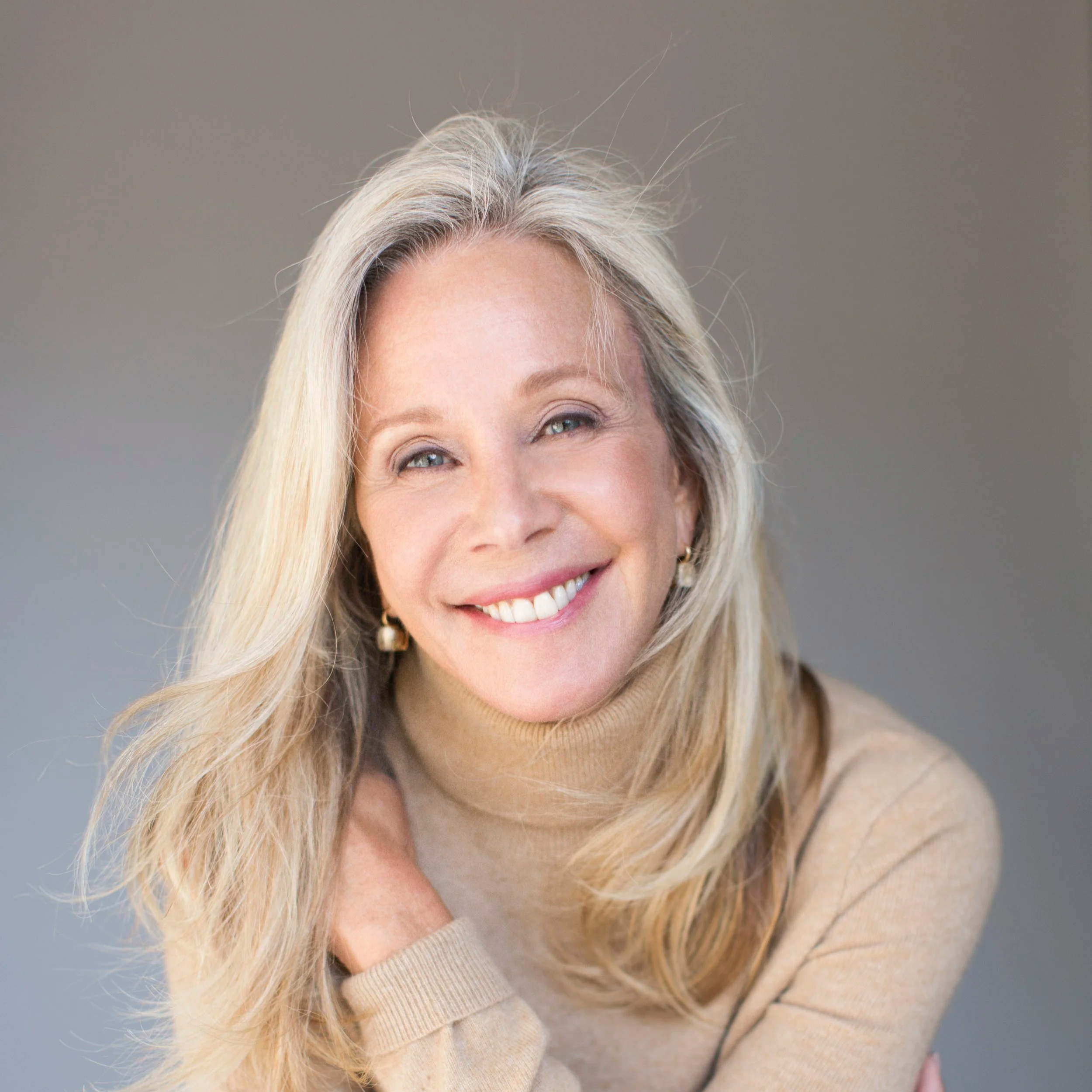 A smiling woman with long blonde hair, wearing a beige turtleneck sweater, sitting against a neutral background.