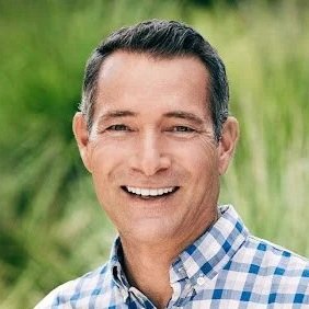 Man smiling outdoors, wearing a blue and white checkered shirt