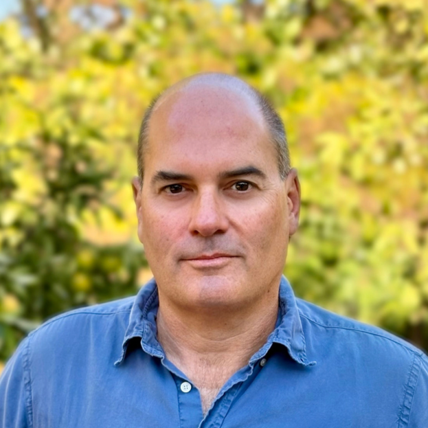 A man with a bald head and light skin, wearing a blue button-up shirt with rolled sleeves, stands outdoors in front of a background of green and yellow foliage.
