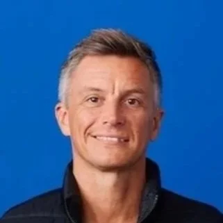 Smiling middle-aged man with short gray hair wearing a black sleeveless jacket over a black shirt, standing against a solid blue background.