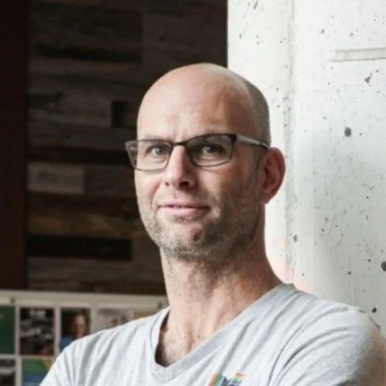 A man with glasses and a light gray t-shirt standing with arms crossed near a concrete wall with a red marking, in an indoor setting with wooden wall elements and posters in the background.