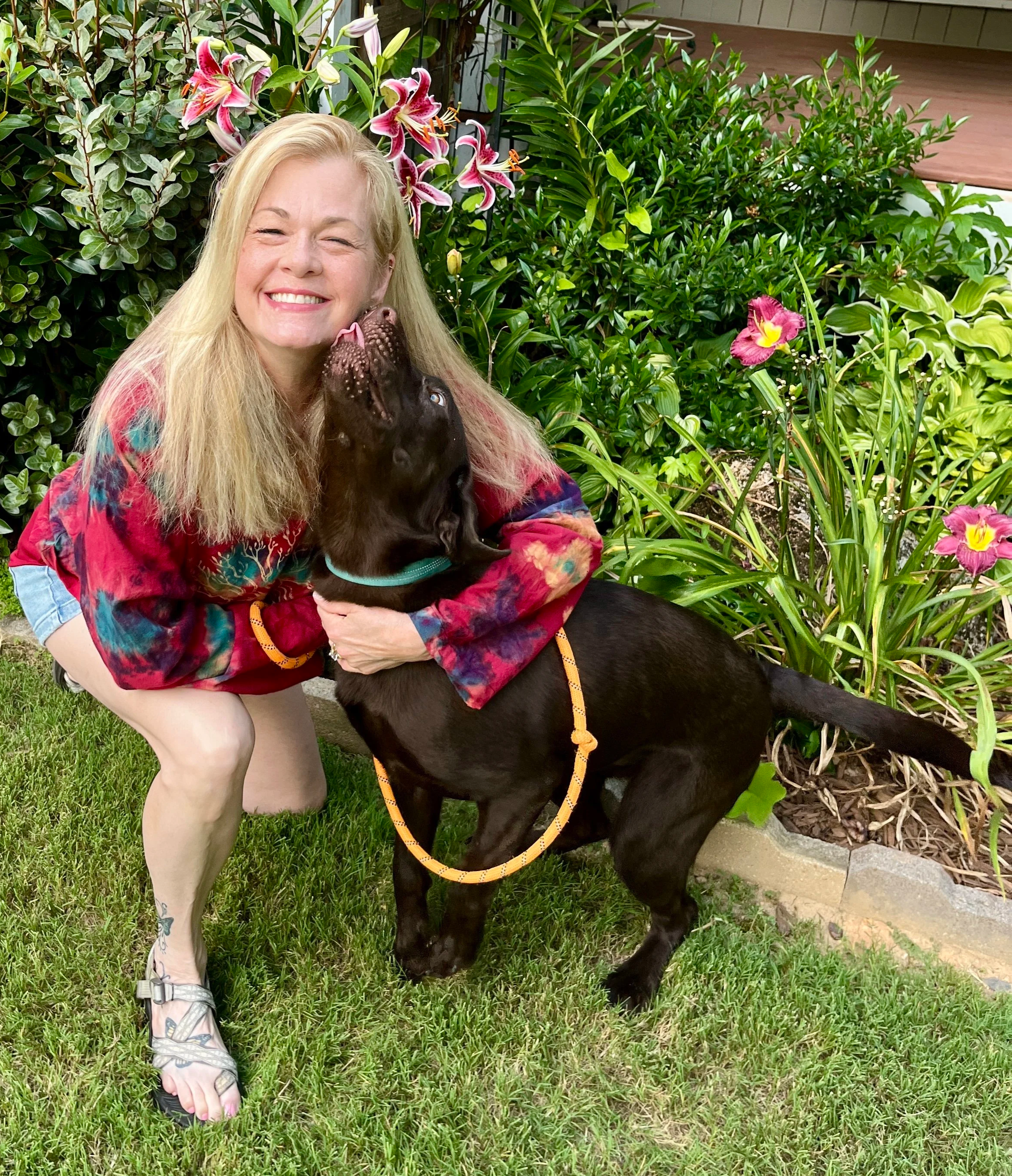 A smiling blonde woman in a colorful tie-dye shirt crouches on the grass, hugging a black Labrador retriever puppy with a purple flower in its mouth. She is wearing a yellow bracelet and silver sandals. The background features lush green plants and pink lilies.