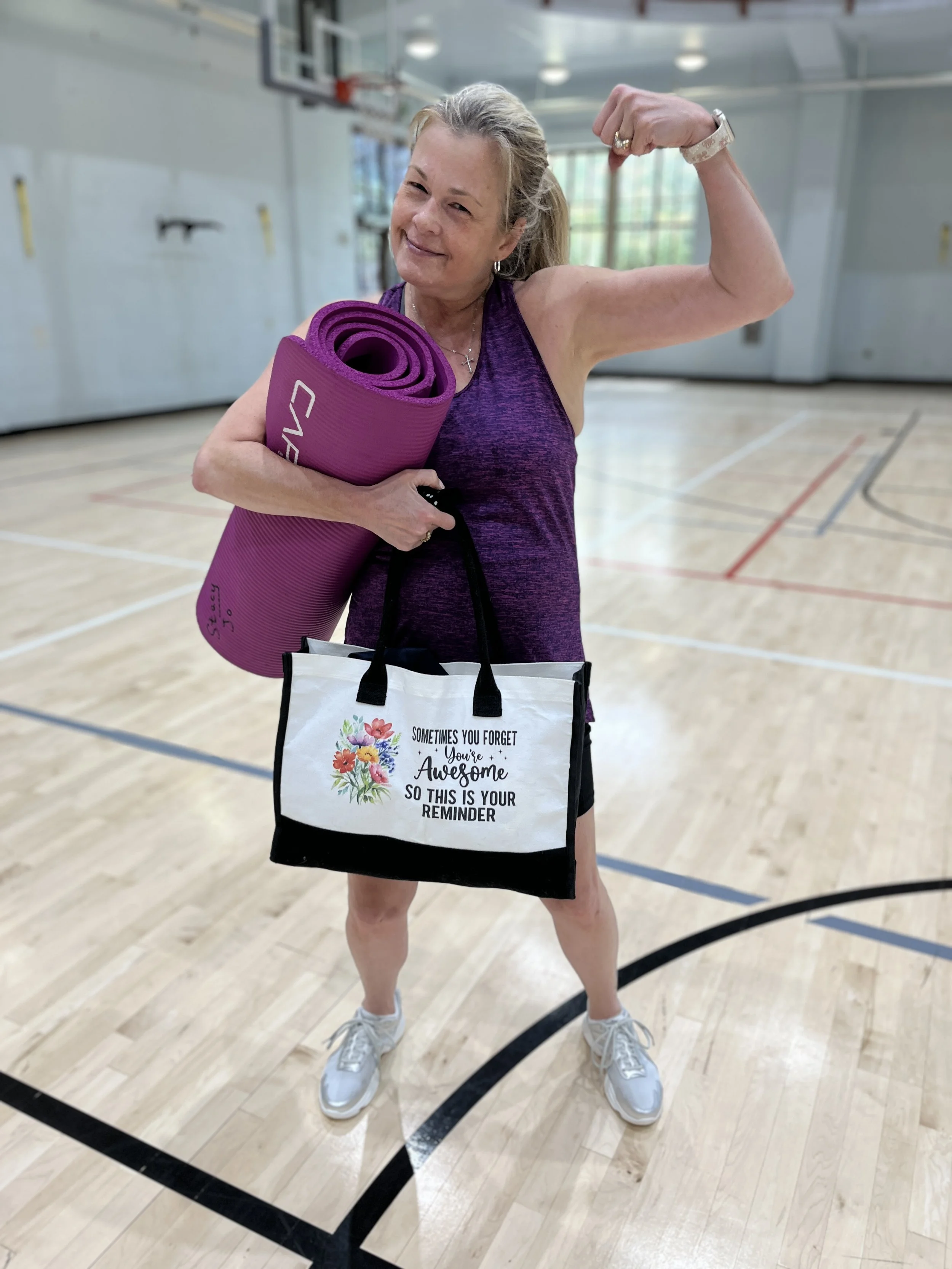 Hippie Chick is flexing her right arm while holding a purple yoga mat in her left arm, standing in an indoor gymnasium. She is smiling.