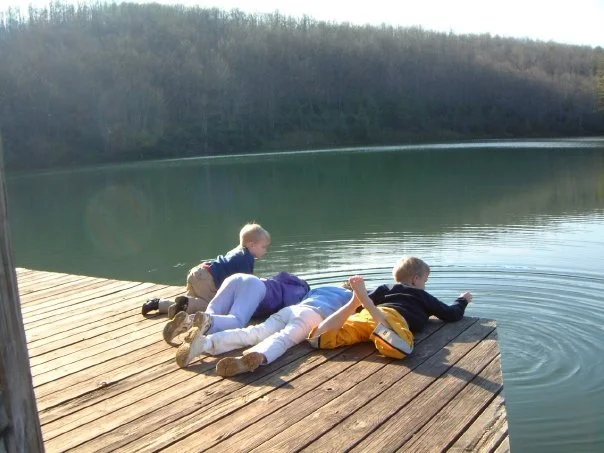 Three children lying on a wooden dock by a lake, with two of them leaning over the water and one looking into the water.