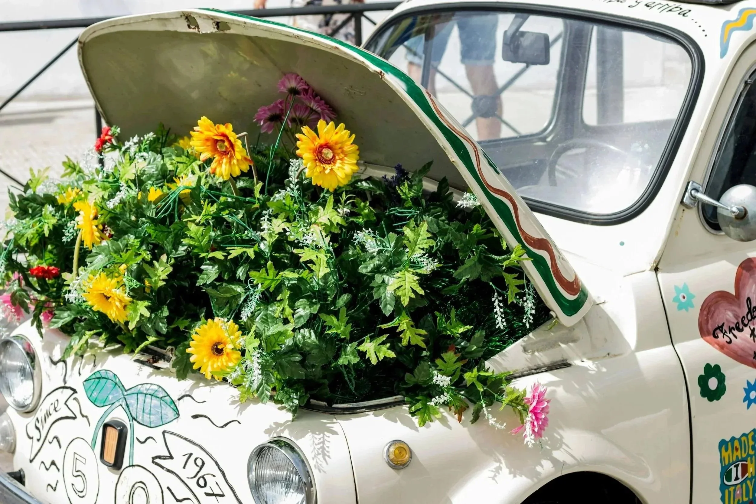 An old white vintage car with its hood open, filled with colorful artificial flowers and decorated with stickers and drawings.