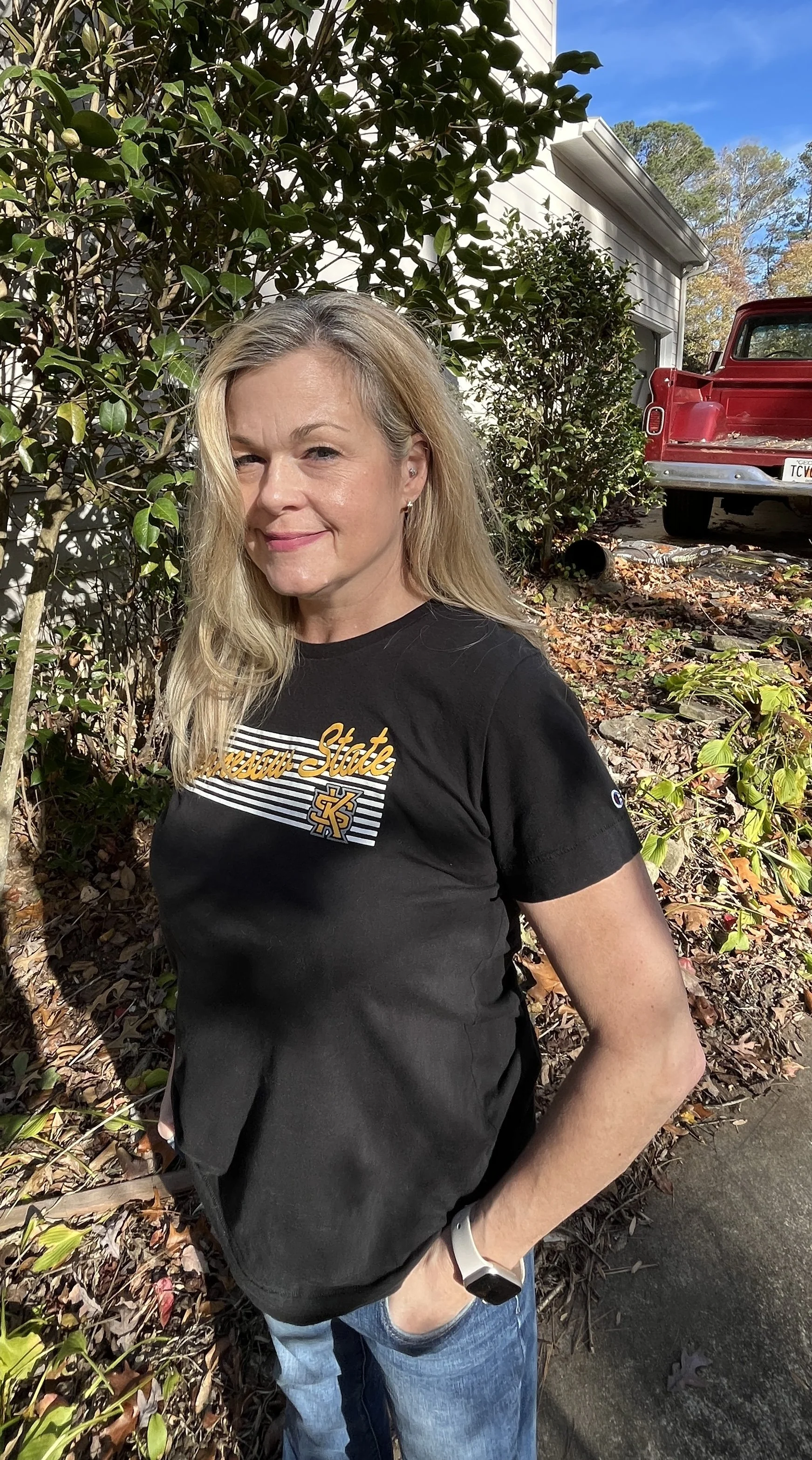A woman standing outdoors near bushes, wearing a black T-shirt with the 'Kennesaw State' logo, blue jeans, and a smartwatch, with a red 1965 truck and a house in the background.