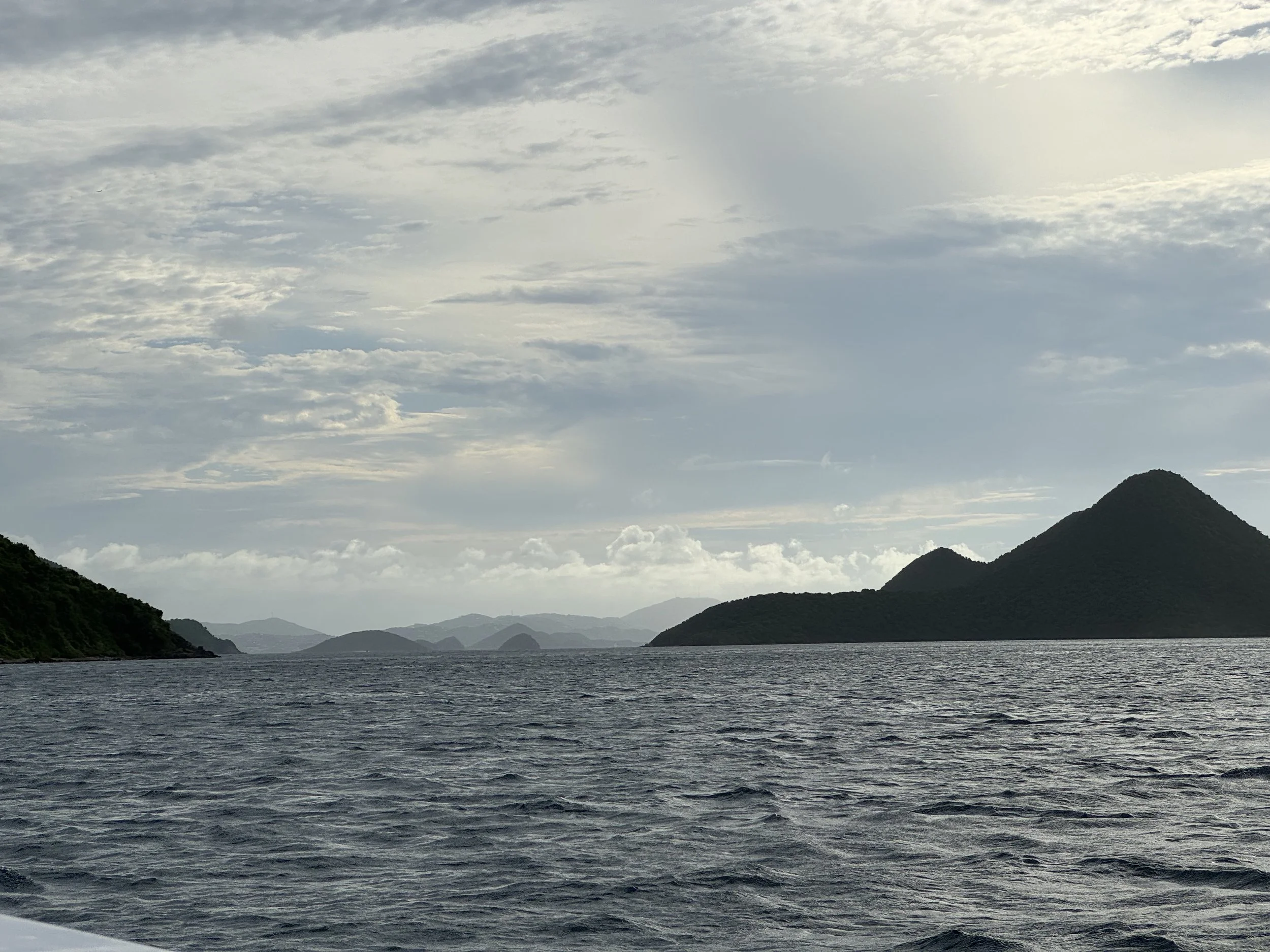 Ocean bay with small waves, surrounded by distant lush green islands and hills under a partly cloudy sky.