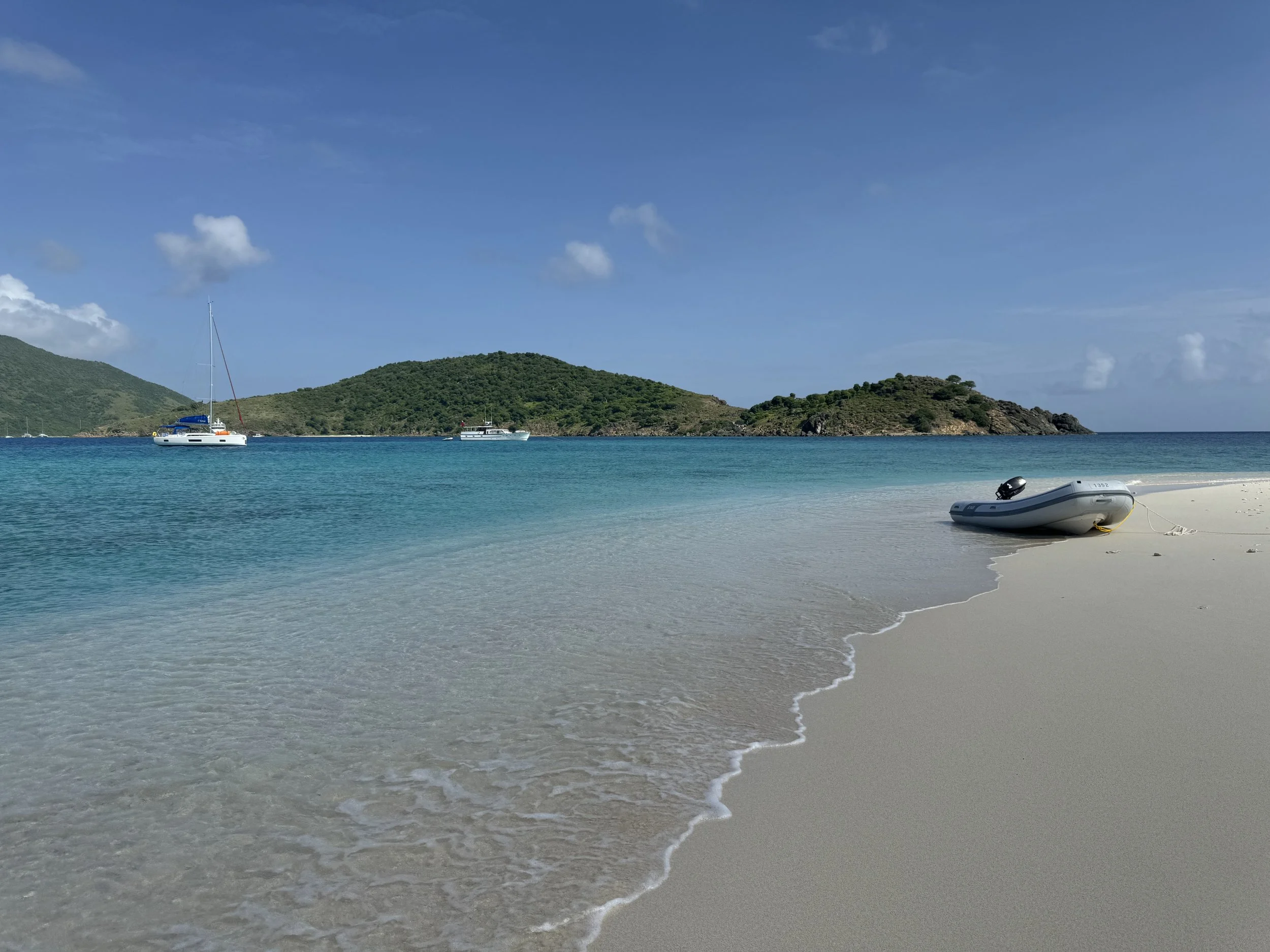 A boat on a sandy beach with turquoise water, green hills in the background, and a clear blue sky.