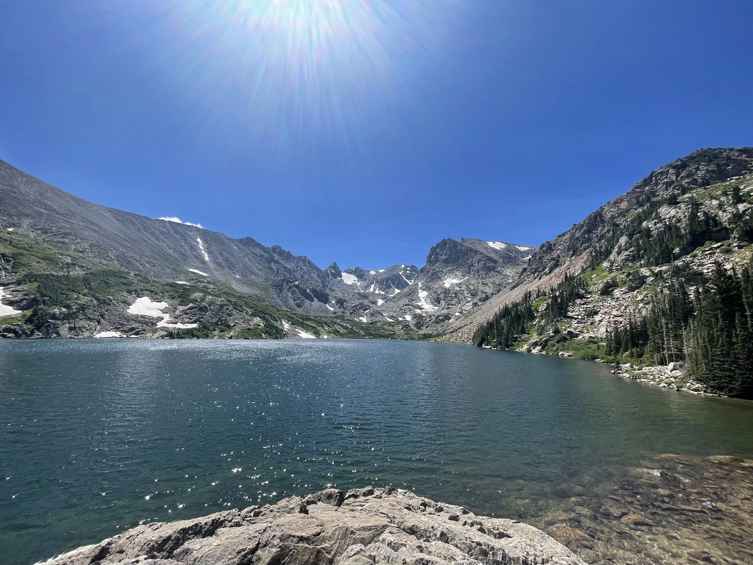 A mountain lake on a sunny day with clear blue skies, surrounded by rocky mountains with patches of snow, and evergreen trees on the right side.