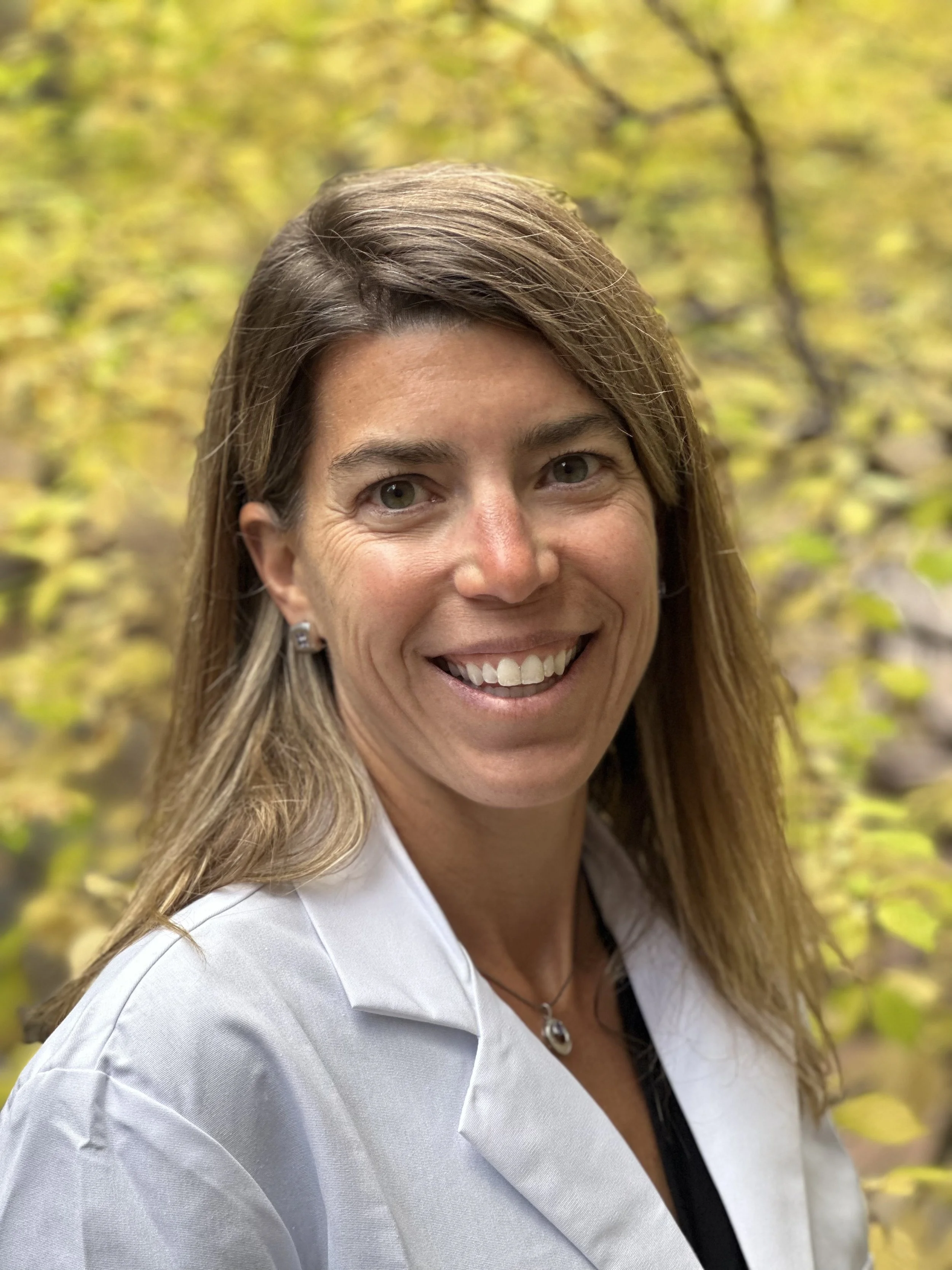 A woman smiling outdoors with blurred yellow leaves in the background, wearing a white coat and a necklace.