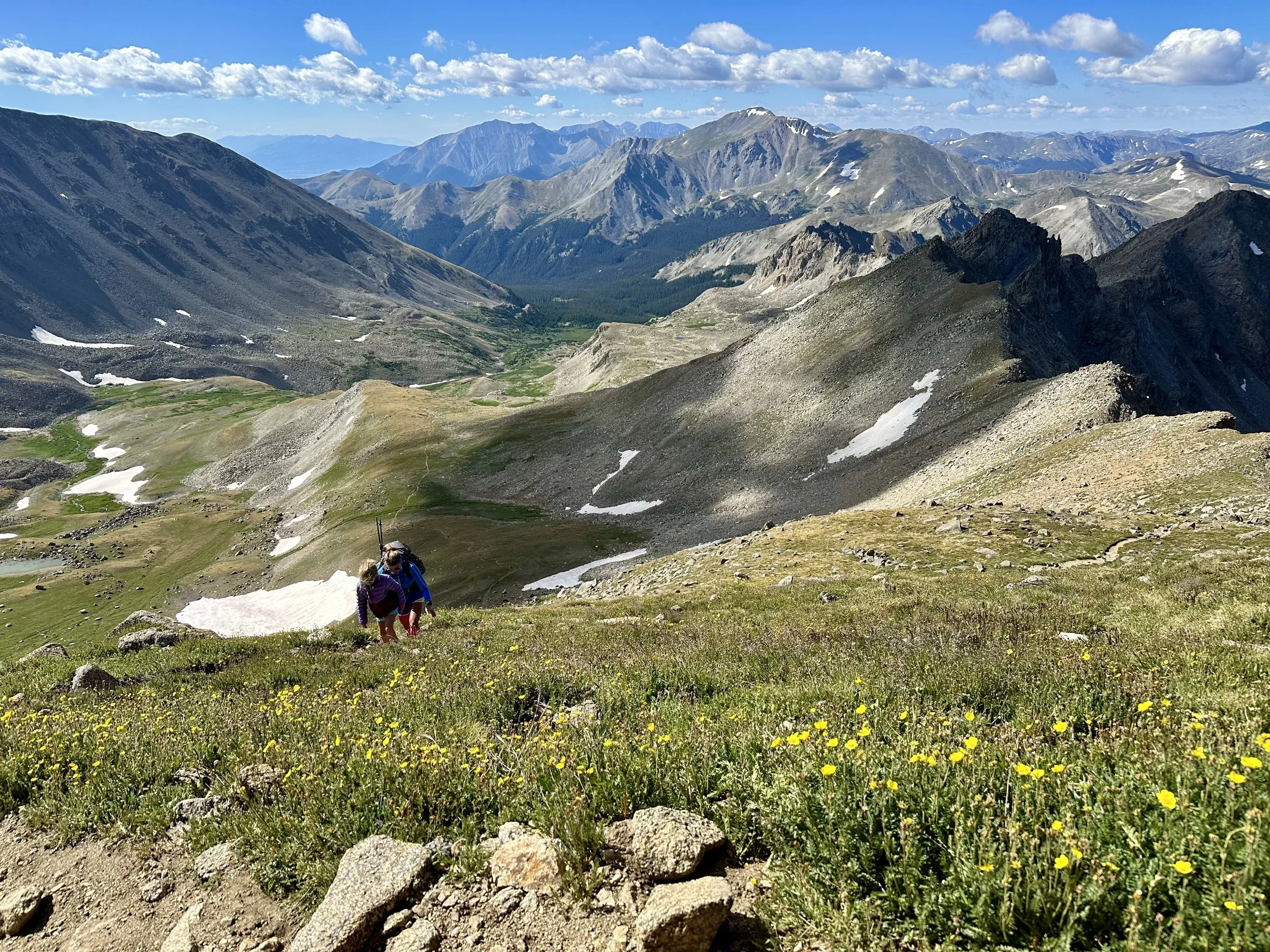 Two hikers climbing a grassy mountain trail with a scenic view of rugged mountains, patches of snow, and a cloudy sky in the background.