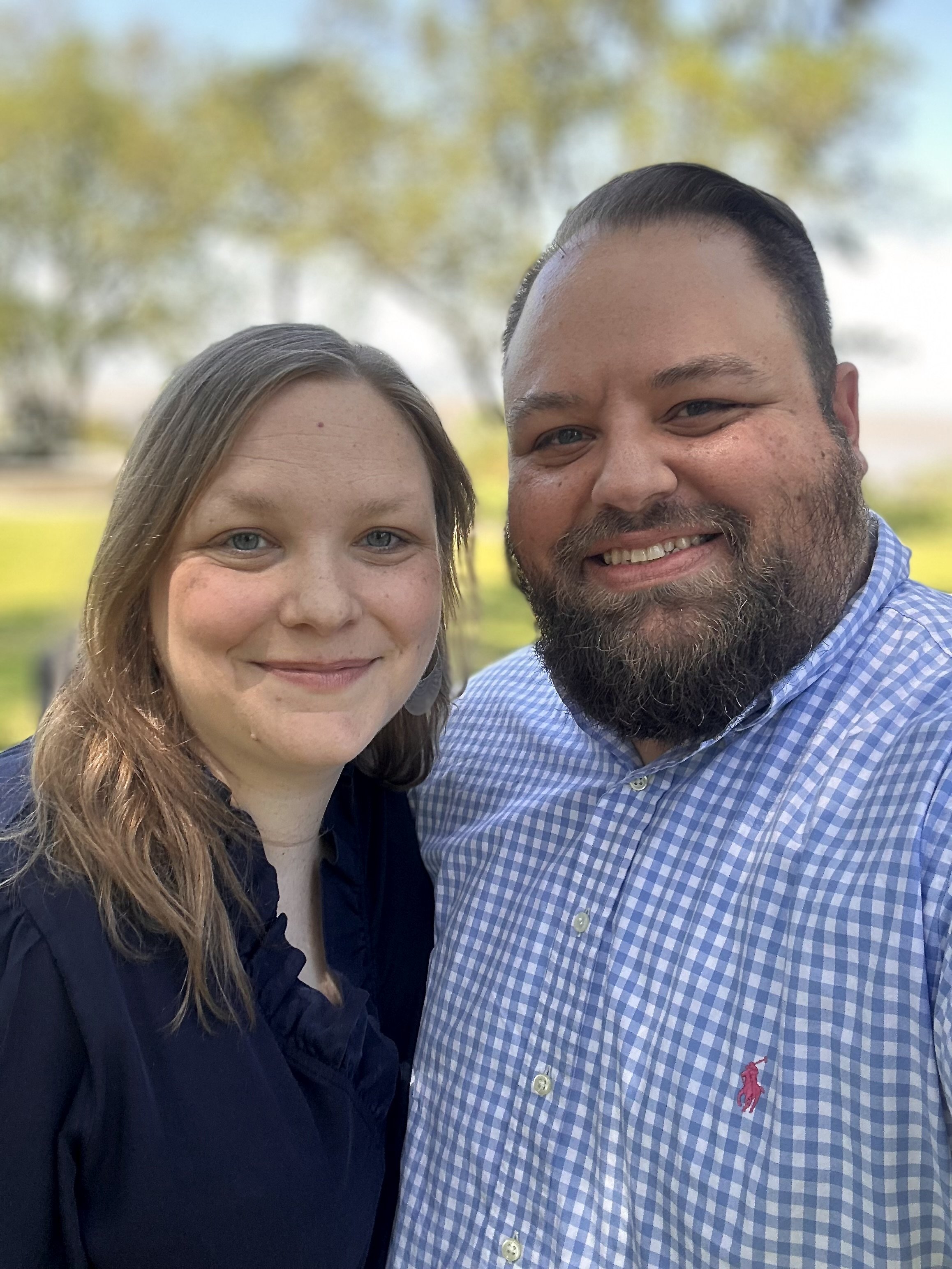 A man and a woman smiling together outdoors with trees in the background.