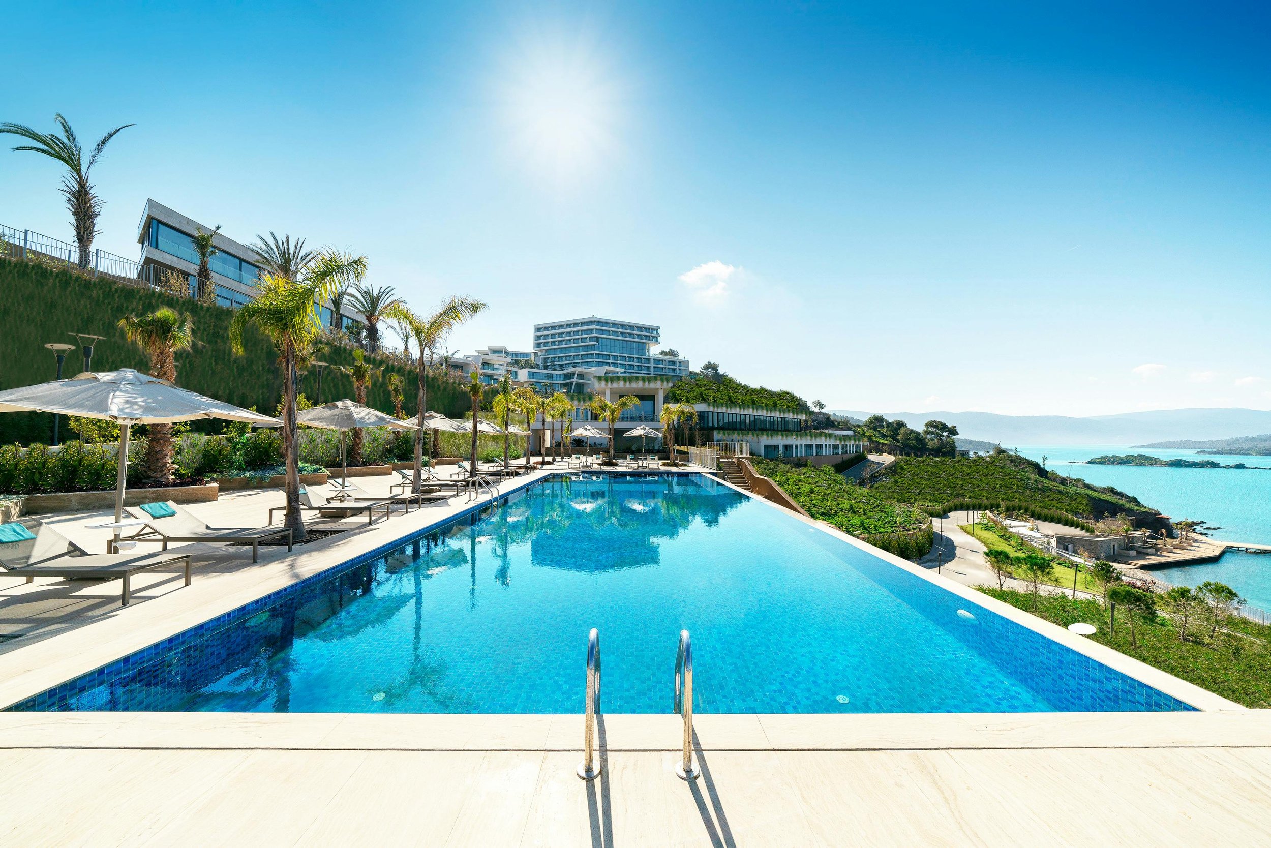Luxury pool with lounge chairs and umbrellas, overlooking ocean and modern buildings on hillside under clear blue sky.