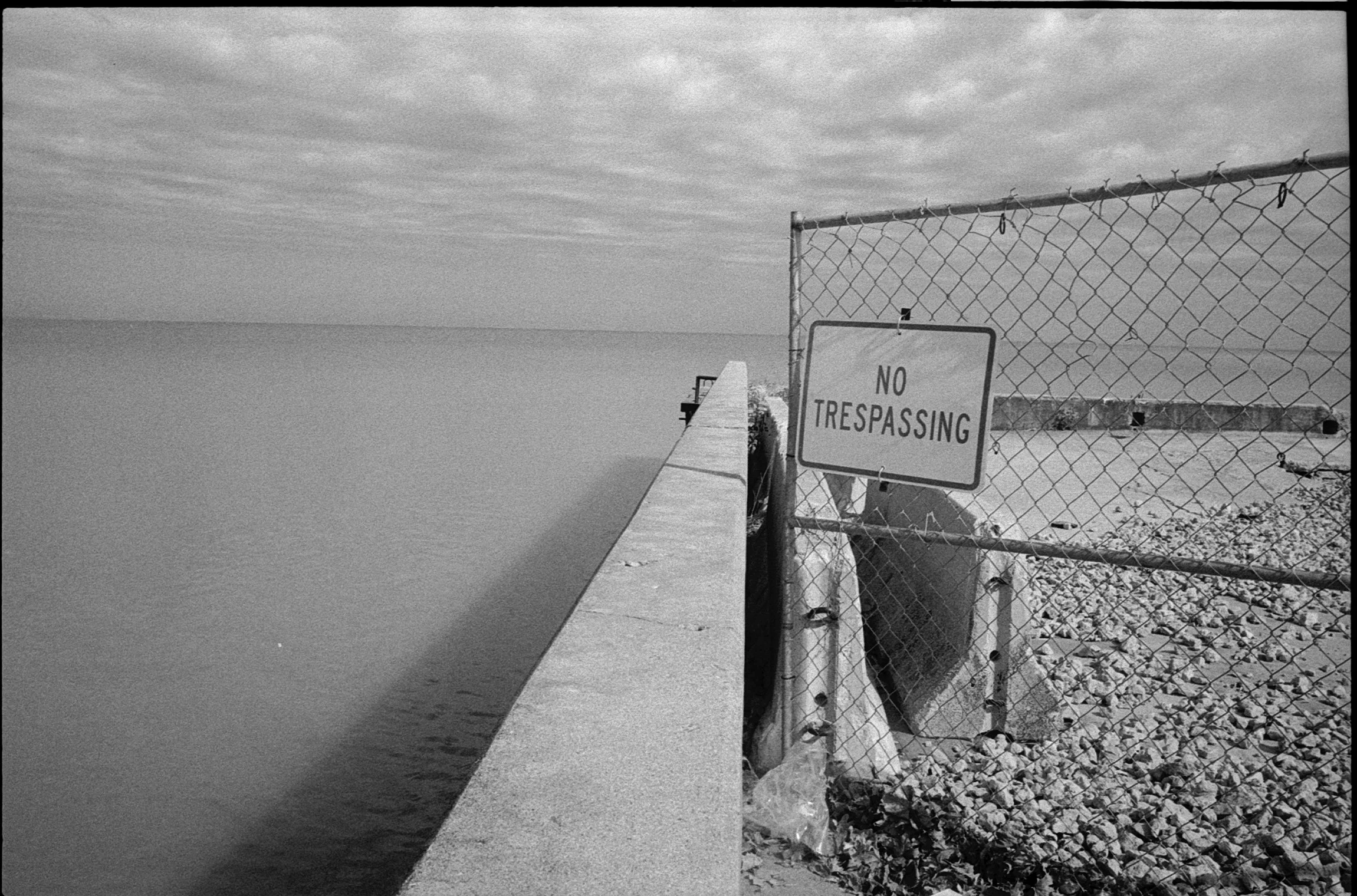 A fence with a 'No Trespassing' sign next to a concrete pier extending into the water, with a cloudy sky overhead.