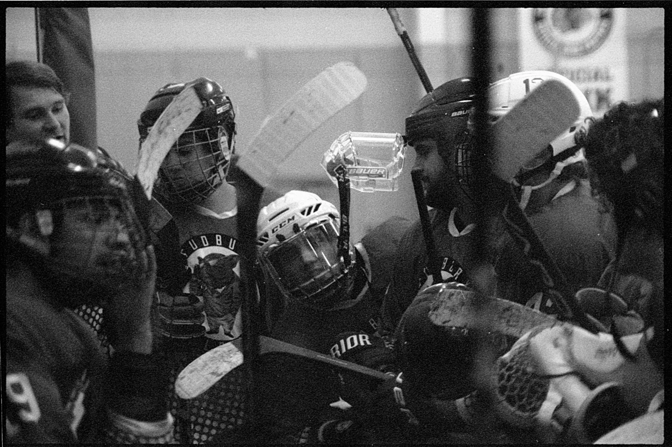 Hockey team huddled together during a game, wearing helmets and gear, with one player raising his arm and holding a water bottle.