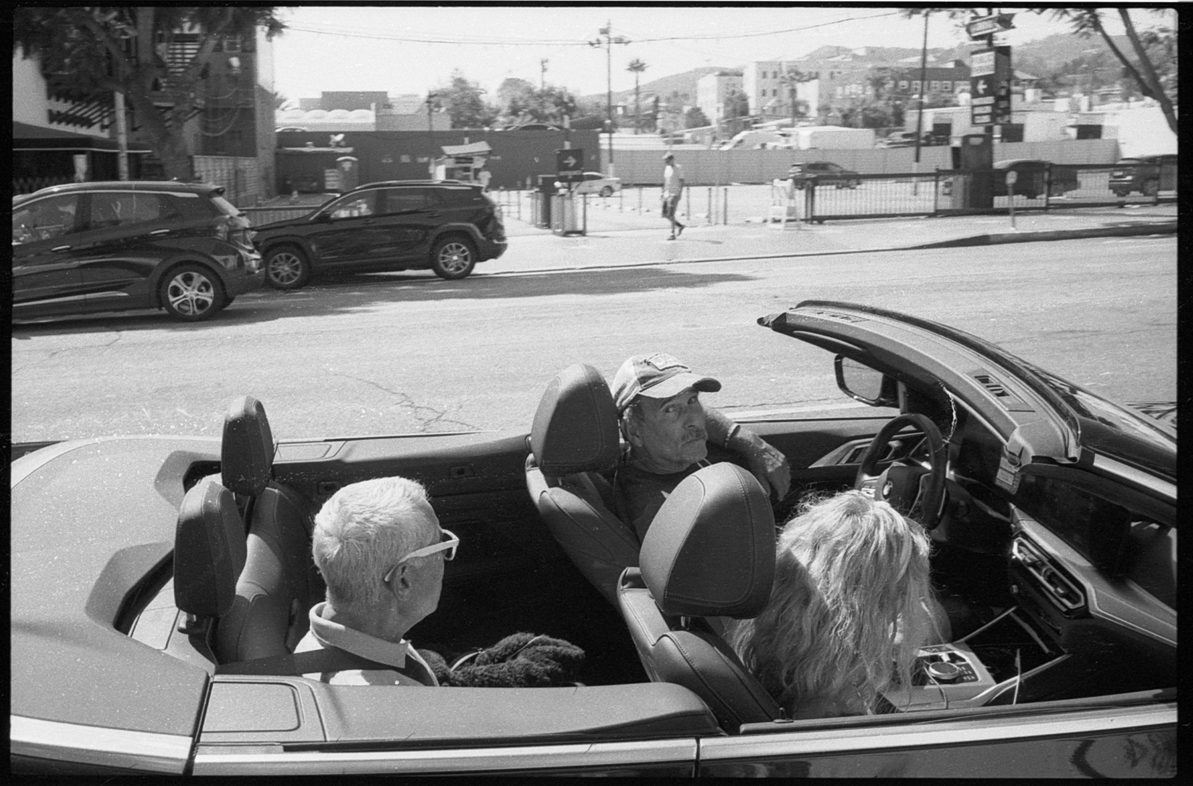 Black and white photo of three people sitting in a convertible car parked on the street. One man, wearing a cap, is in the back seat looking out. A woman with curly hair, wearing glasses, is in the front passenger seat looking at something outside th