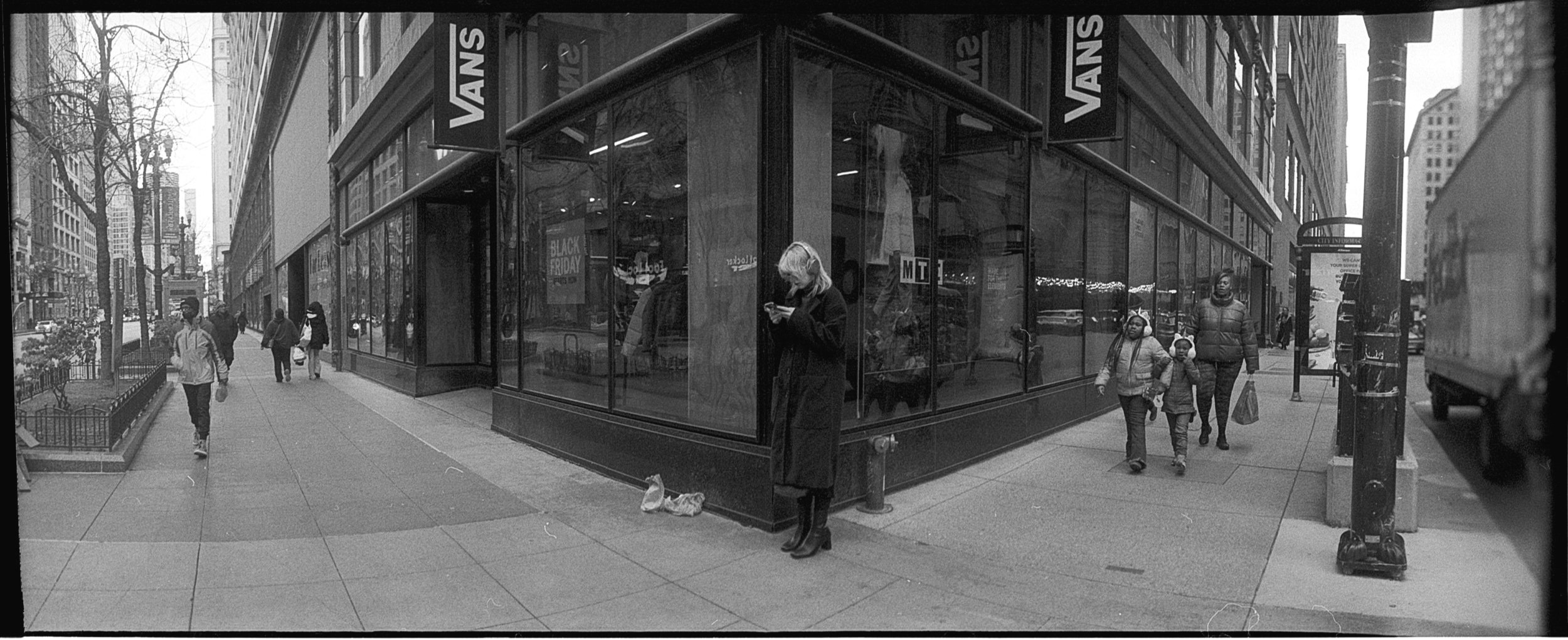 Black and white photo of a city sidewalk with pedestrians walking past a storefront with large glass windows. A woman is standing at the corner, looking at her phone, with a shopping bag on the ground beside her. Two children and an adult are walking