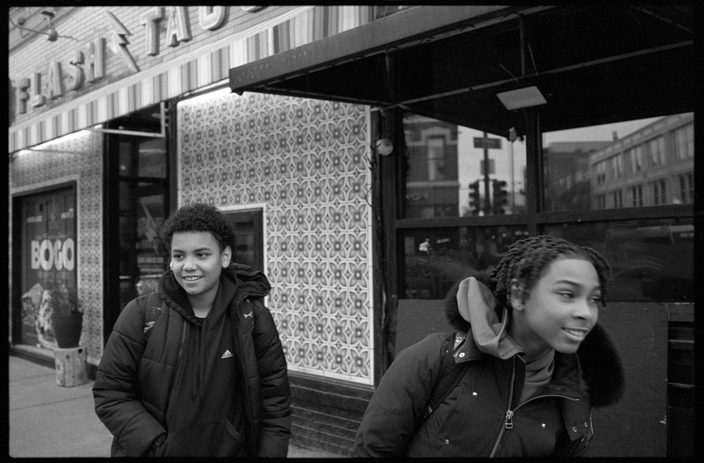 Two young people walking on a sidewalk in front of a building with decorative patterned walls and large windows.