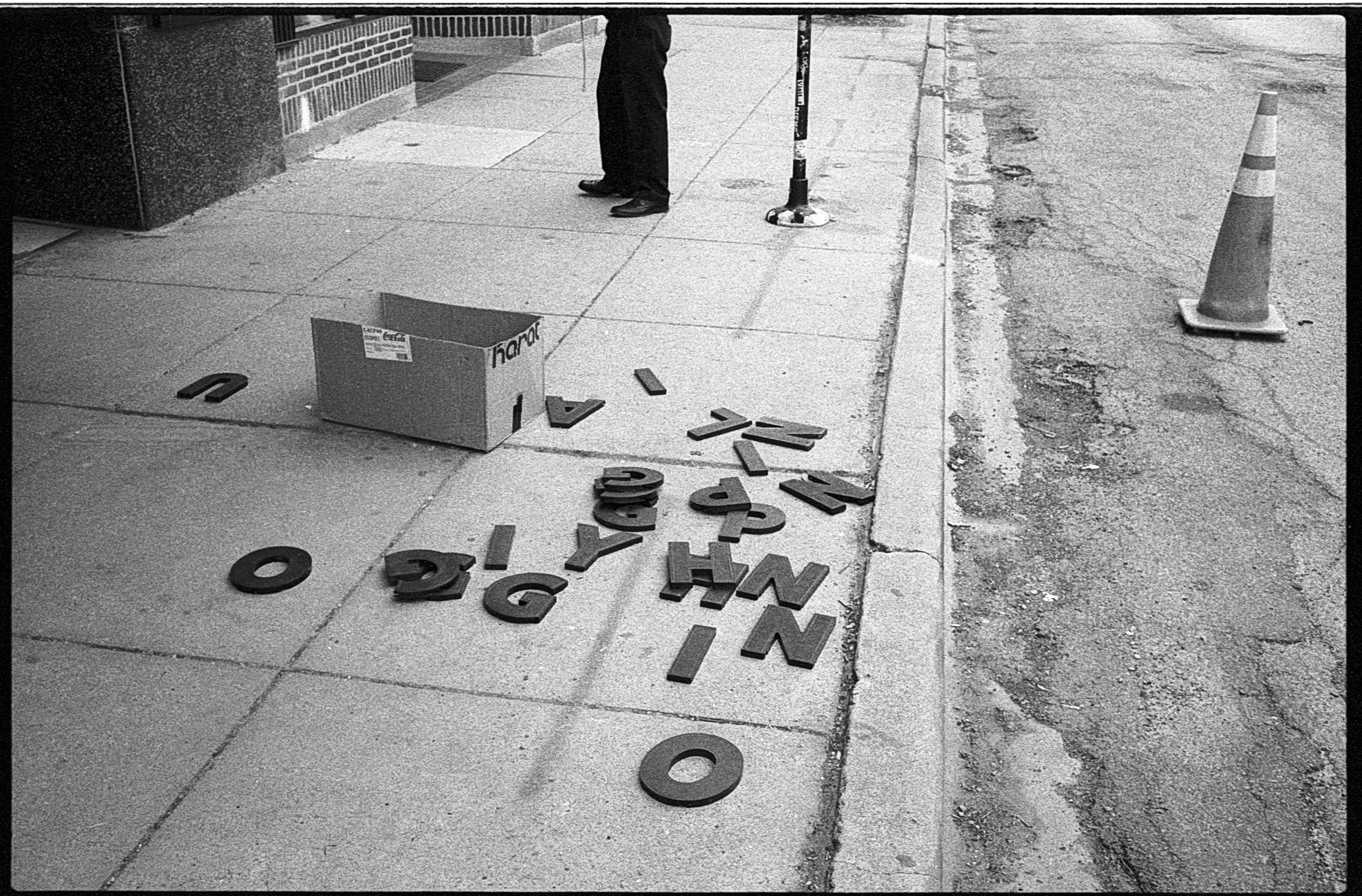 Scattered letters on a sidewalk next to an open box, with a person standing nearby and an orange traffic cone in the background.