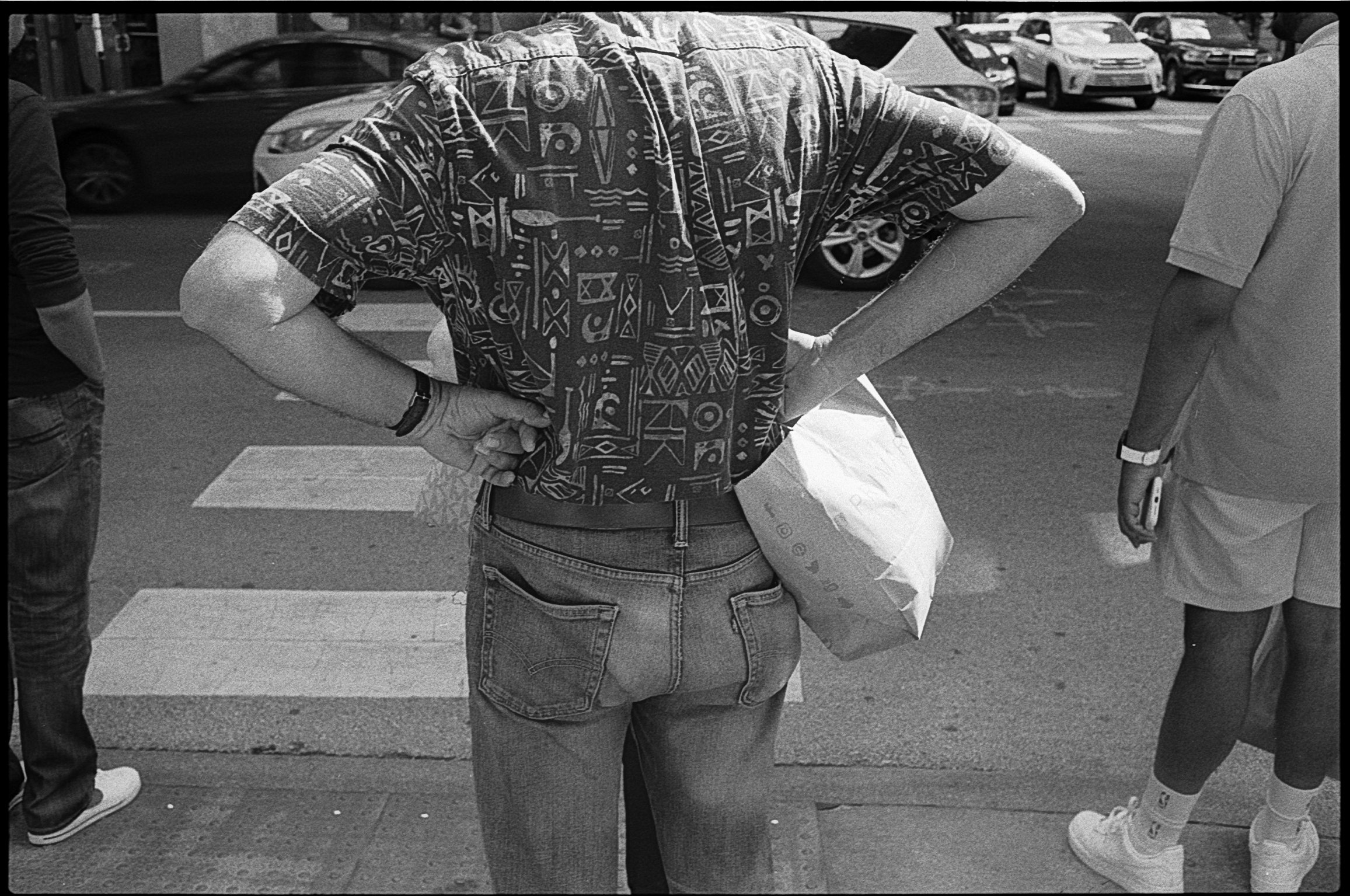 A person wearing a patterned shirt with hands on hips, standing on a street with parked cars and pedestrians visible in the background in black and white