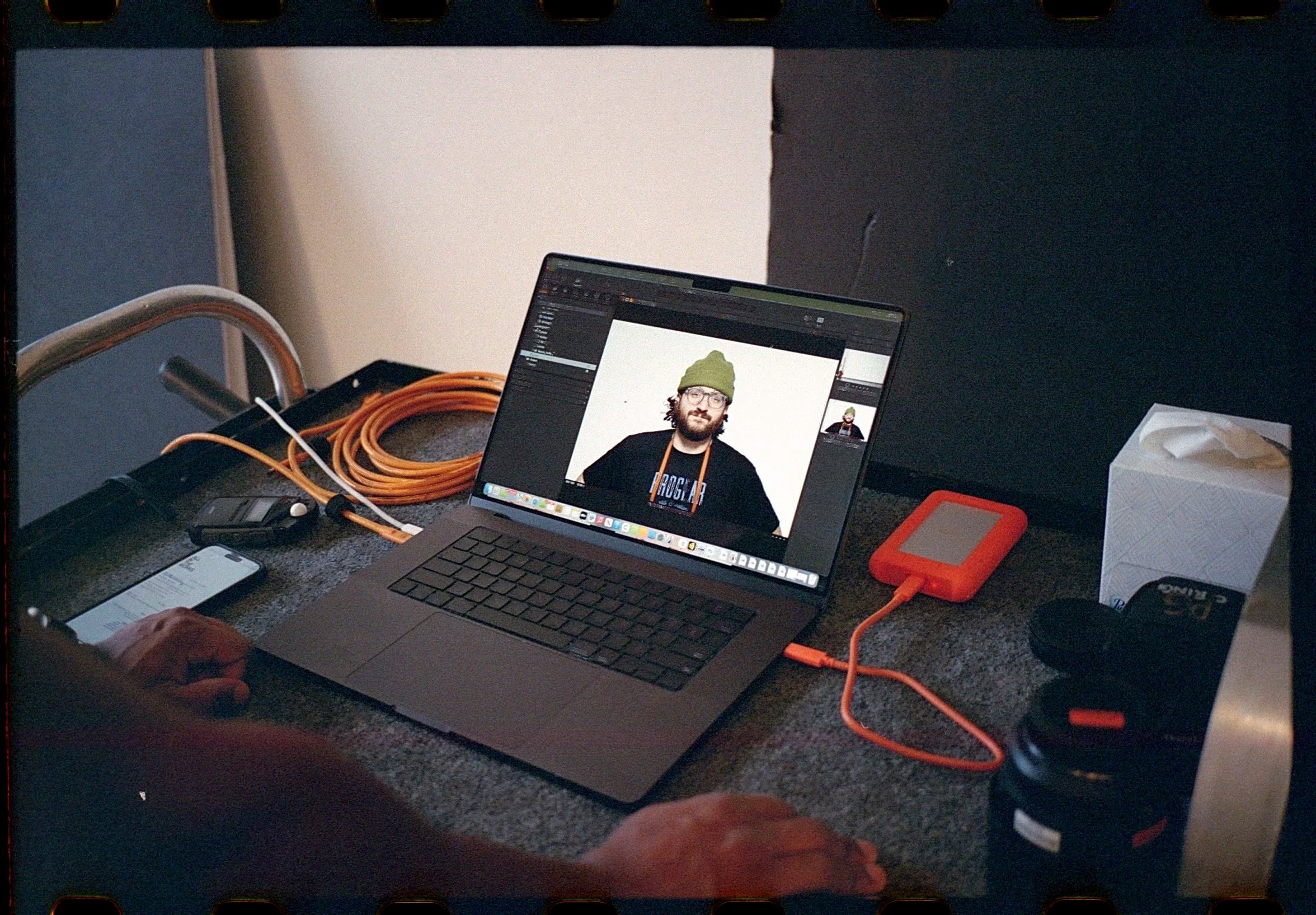 A workspace featuring a laptop displaying a photo editing program with an image of a man wearing a green beanie and glasses. The table has a tissue box, a small external hard drive, a smartphone, an orange extension cord, and headphones. A person's hands are visible, working at the desk.