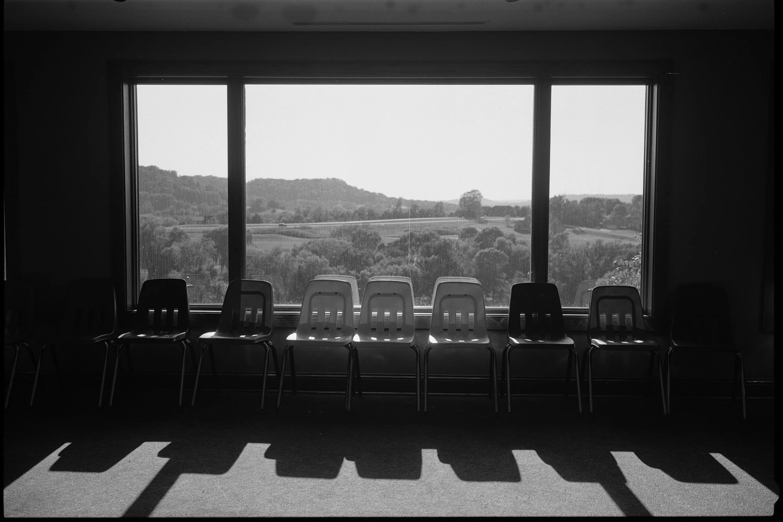 Seating area with nine plastic chairs facing a large window showing a landscape with hills and trees.