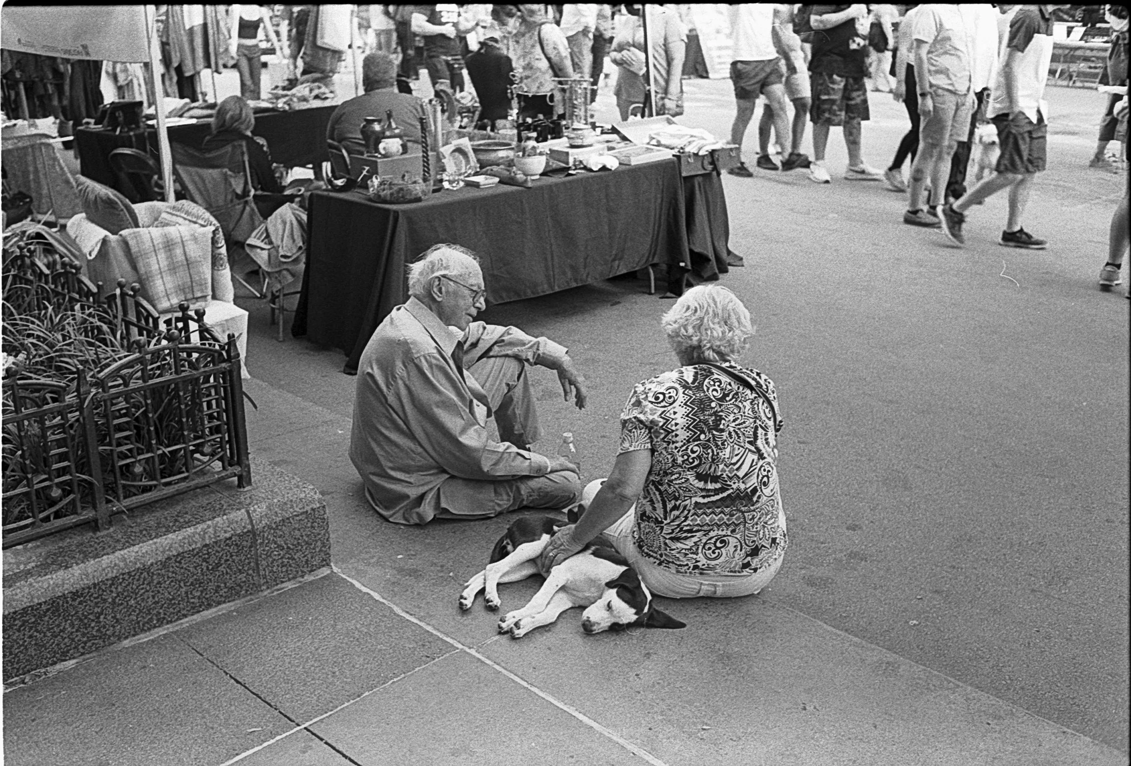 An elderly man and woman sitting on the sidewalk, with the woman petting a dog lying down beside her. A street market is visible behind them, with a table of items and people walking by.