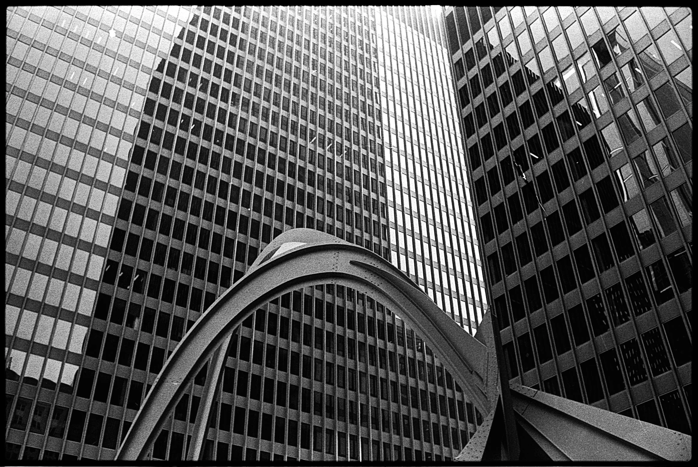 Black and white photo of tall modern skyscrapers with glass windows and reflective surfaces, taken from below, with a curved architectural structure in the foreground.