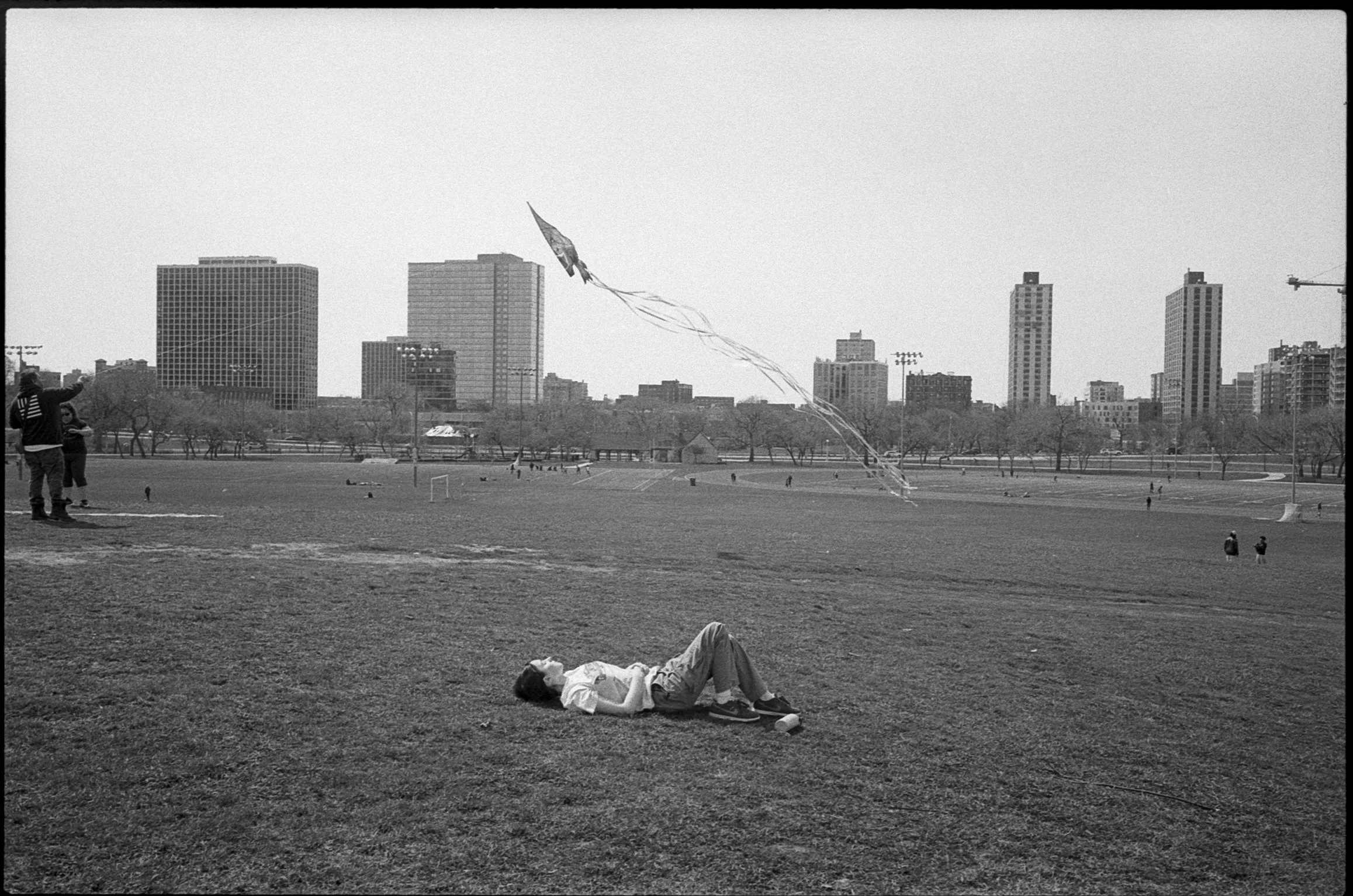 Child lying on grass in a park flying a kite with tall buildings in the background.