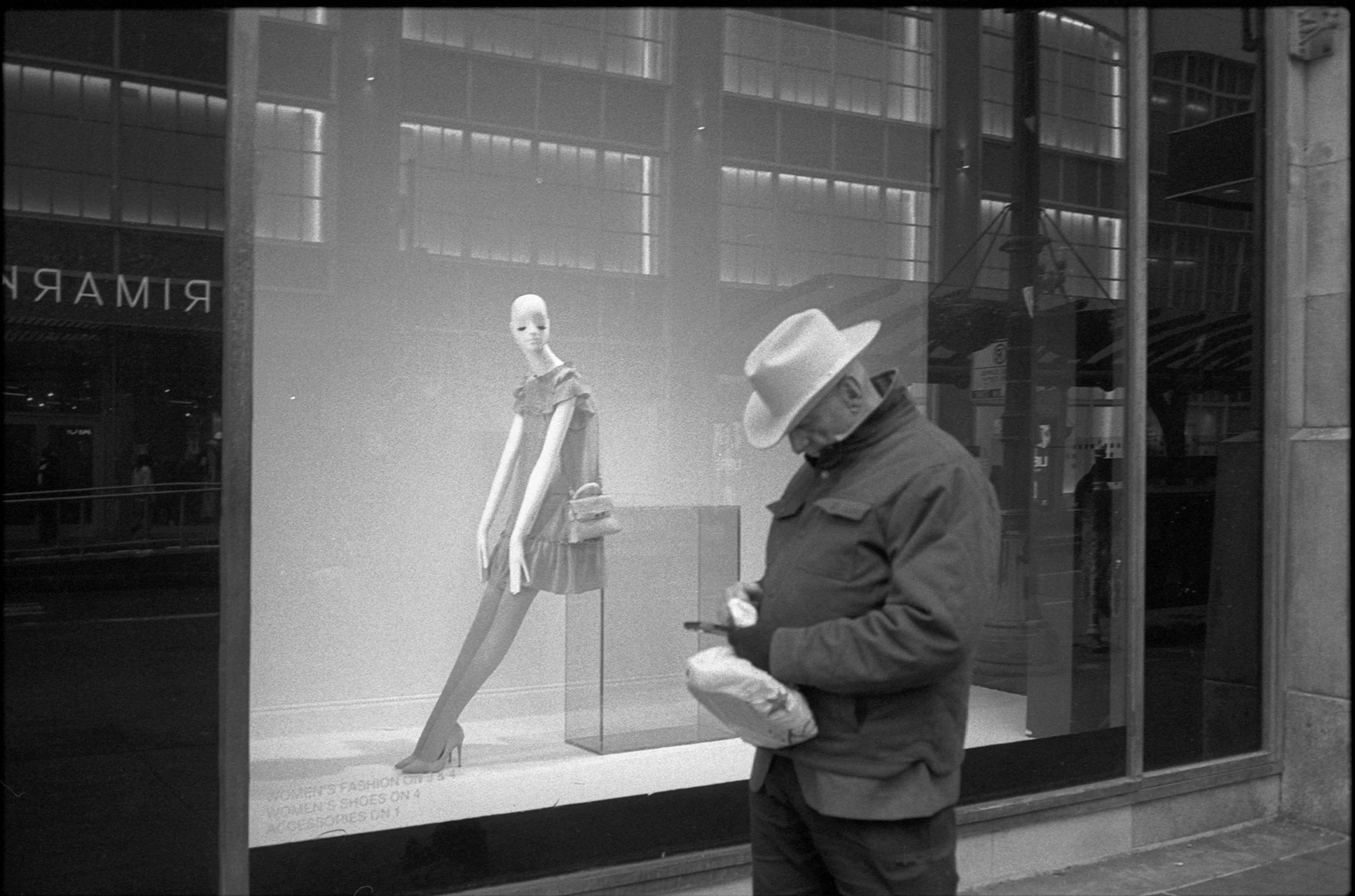 A person wearing a hat and jacket looking at their phone in front of a store window. The window displays a mannequin dressed in a dress with a handbag.