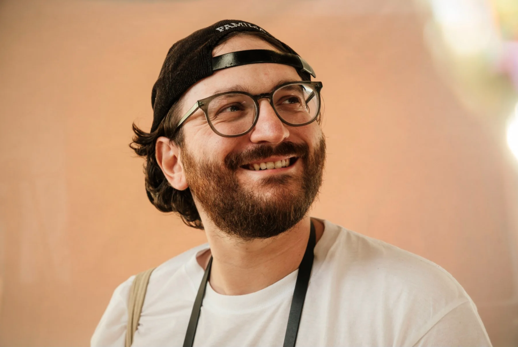 A smiling man with glasses, a beard, and a mustache, wearing a black cap backwards and a white t-shirt, against a light-colored background.