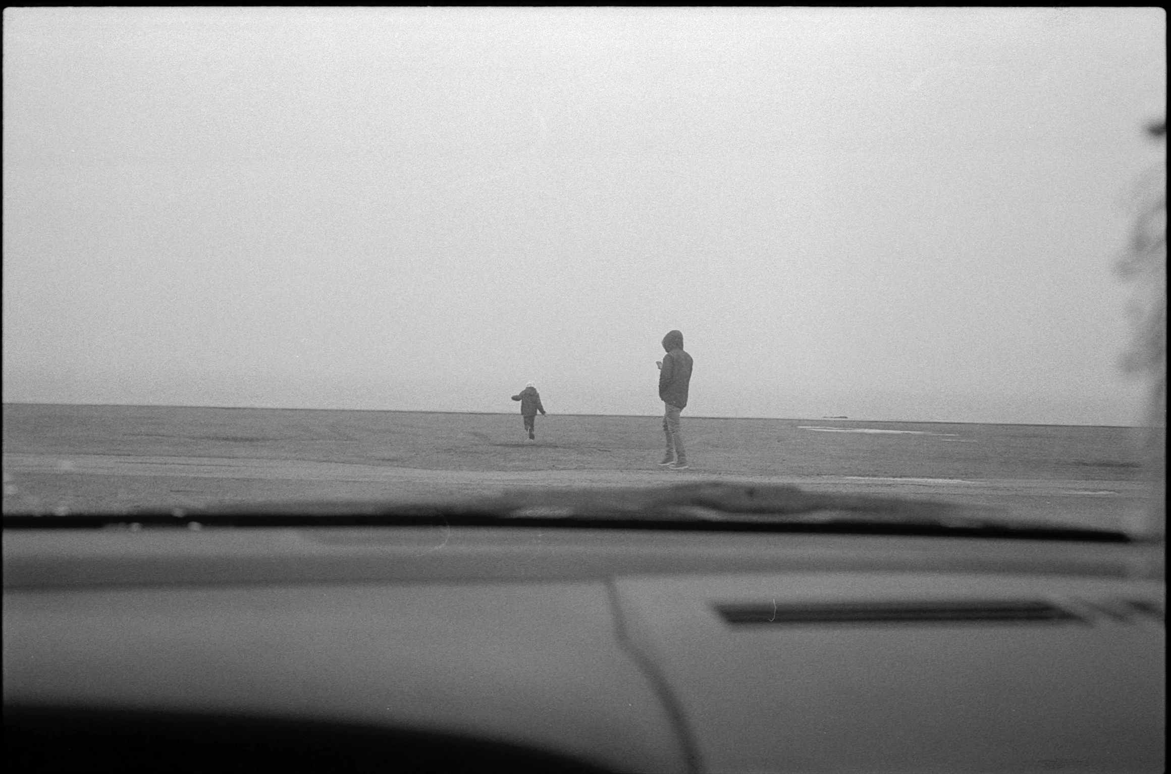 Black and white photo of a person and a child standing on a beach, viewed from inside a car.