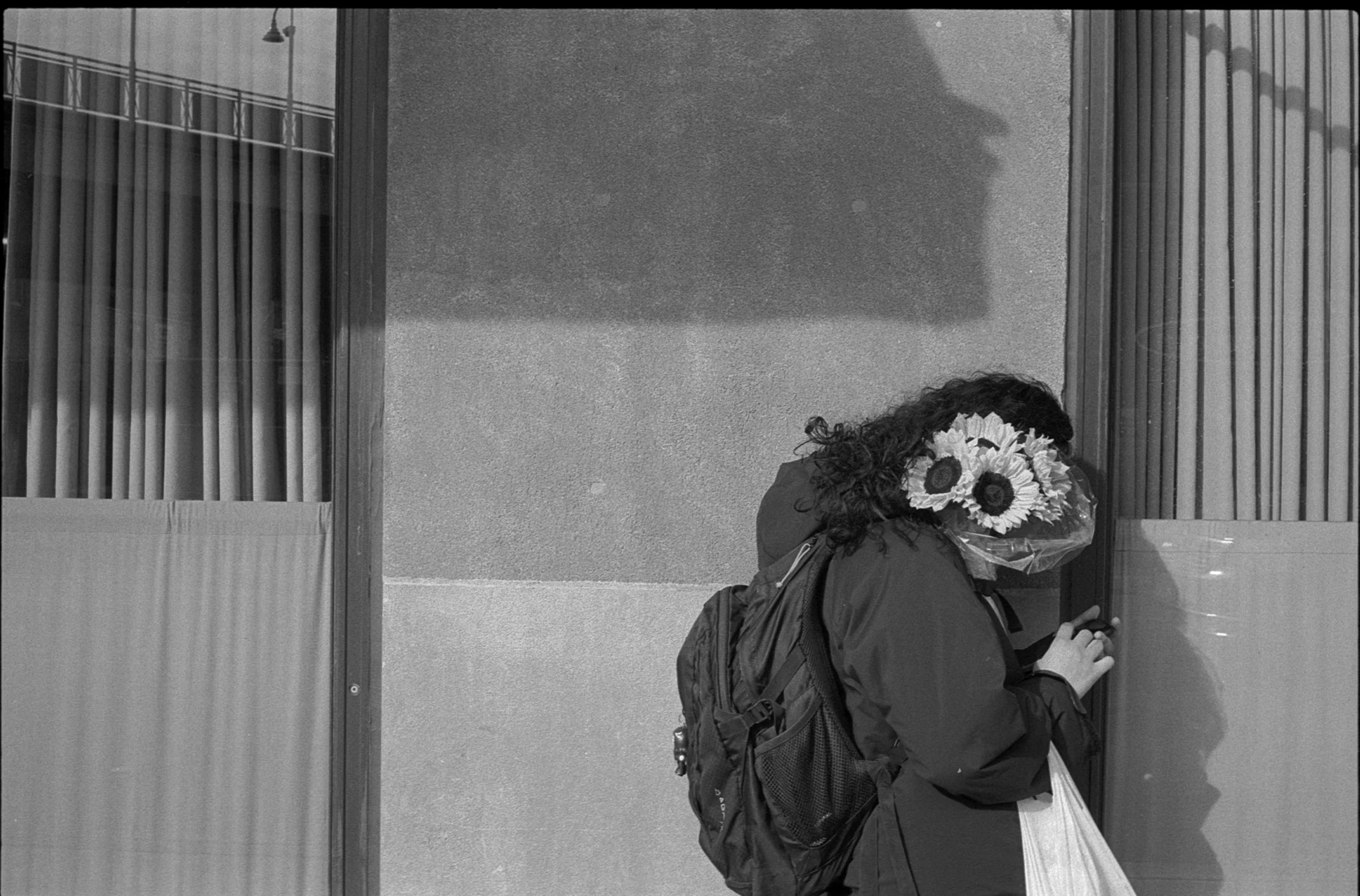 A woman with curly hair is standing outside near a building, holding a bouquet of sunflowers wrapped in plastic, and looking at her phone. She is wearing a backpack and a jacket.