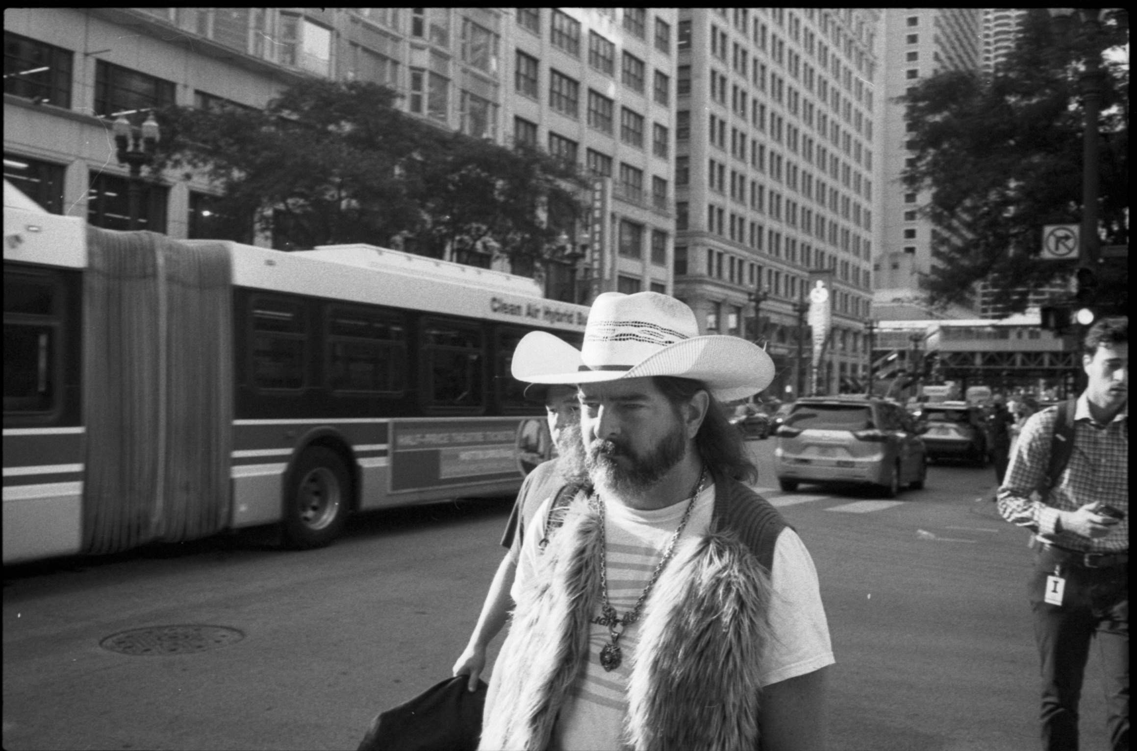 A man wearing a cowboy hat and a fur vest walks on a busy city street with tall buildings, a city bus, and other pedestrians visible in the background.