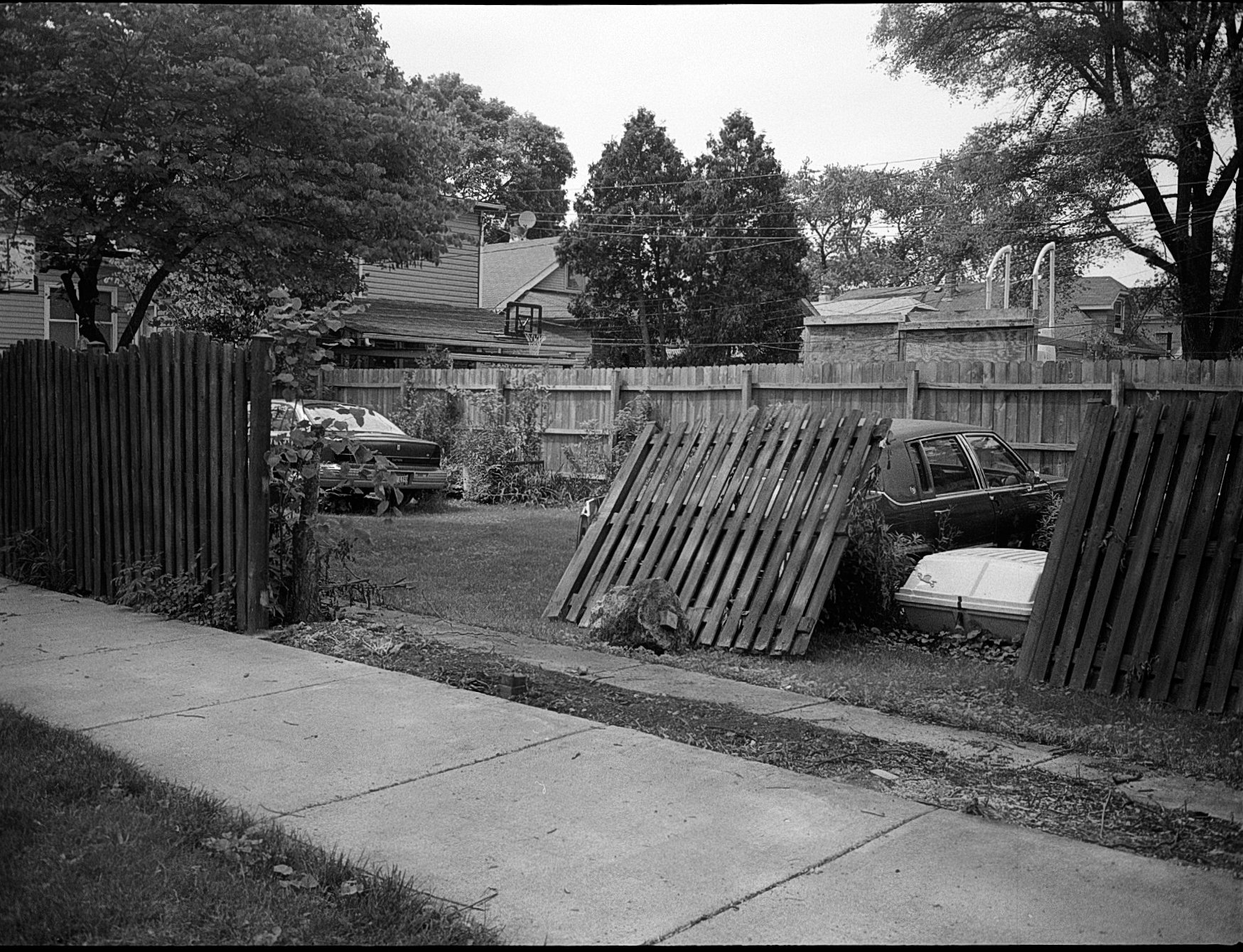 A backyard with a wooden fence, leaning fence sections, two cars, a boat, trees, and a sidewalk in a neighborhood.
