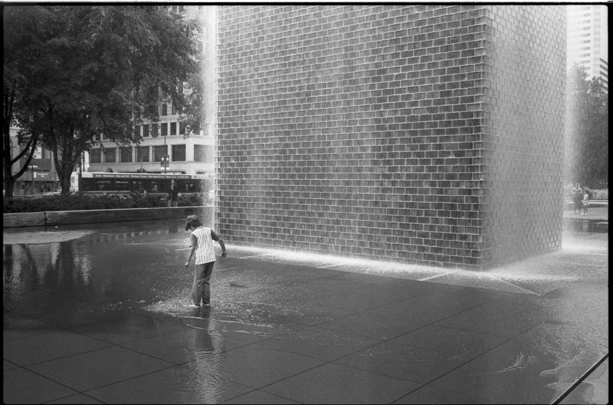 A young boy playing in water at a fountain with a large brick wall in the background in an urban park.