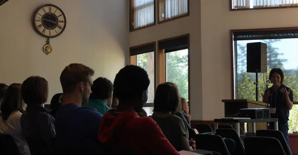 A woman speaking to an audience in a room with large windows and a wall clock.