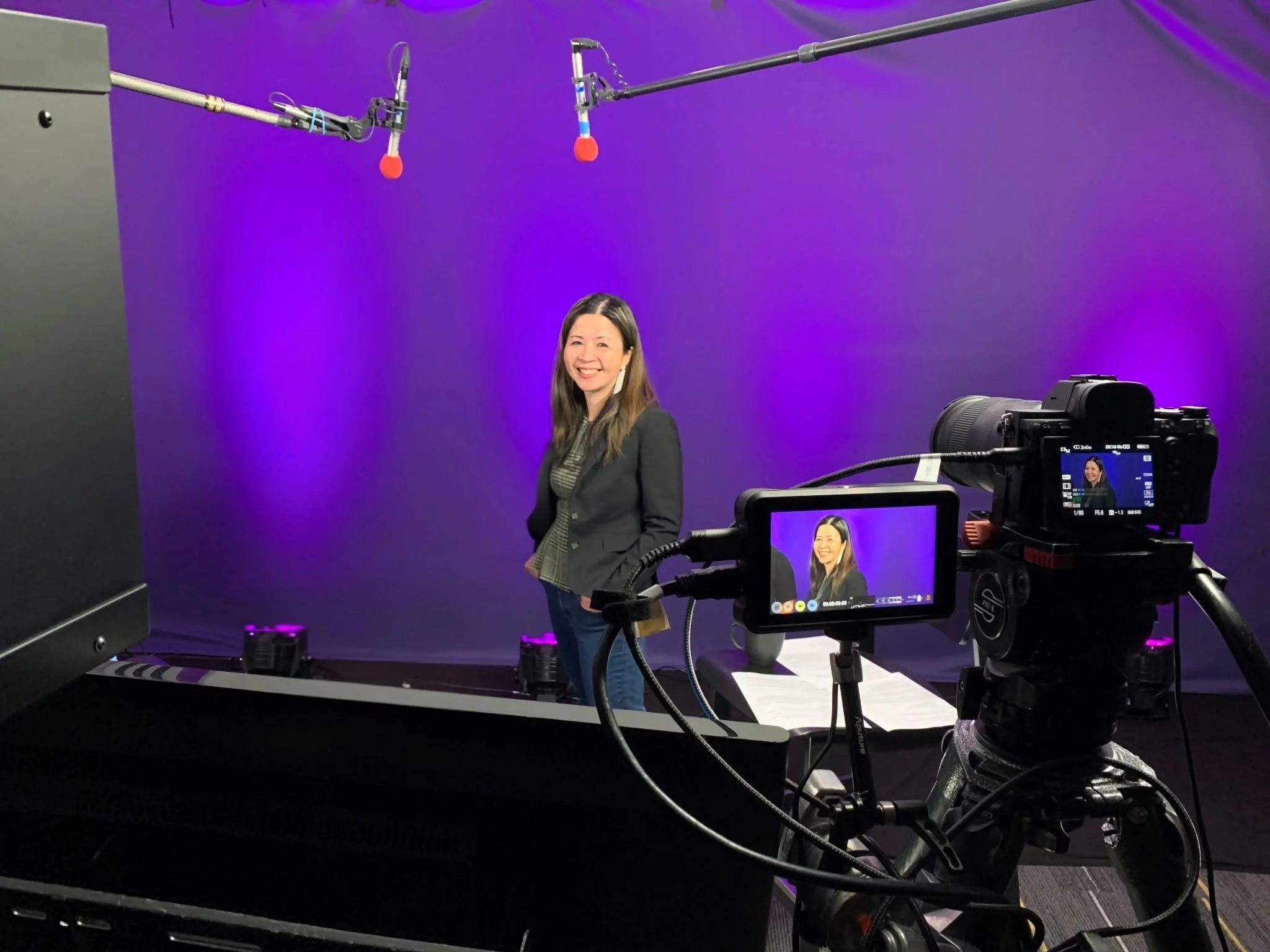 dark haired Asian female in a studio with microphones hanging from the ceiling amidst a purple background.