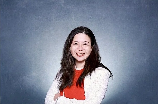 A woman with long dark hair smiling, wearing a red top and a white sweater, standing against a blue-gray gradient background.