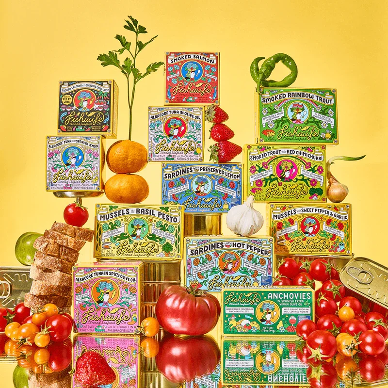 Assorted canned fish products arranged on a yellow background with fresh produce and herbs, including strawberries, cherry tomatoes, a garlic bulb, a tomato, and a sprig of parsley.