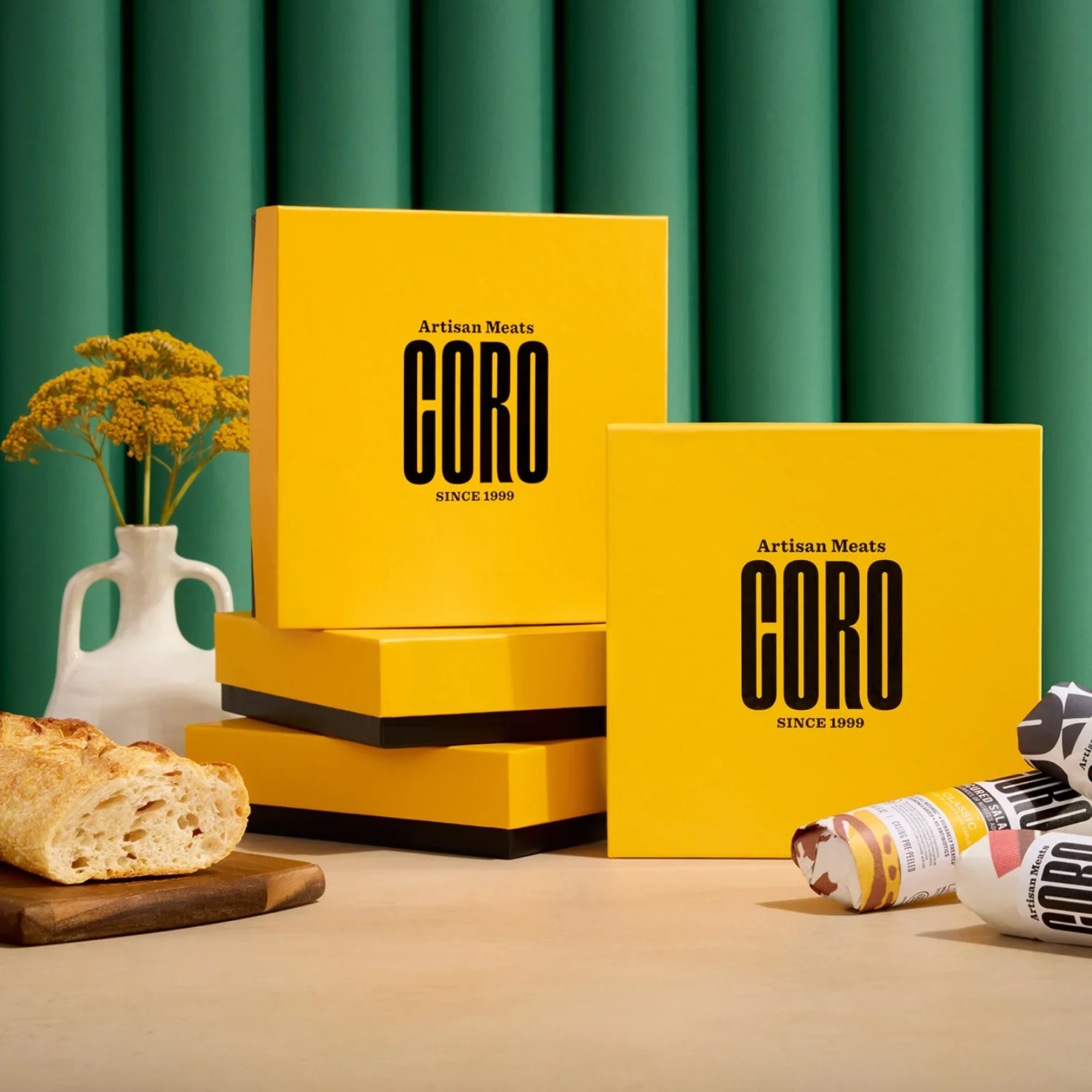 Yellow boxes of Coro artisan meats on a table, with a loaf of bread on a cutting board, a white vase with yellow flowers, green curtains in the background, and some rolled-up packaging.
