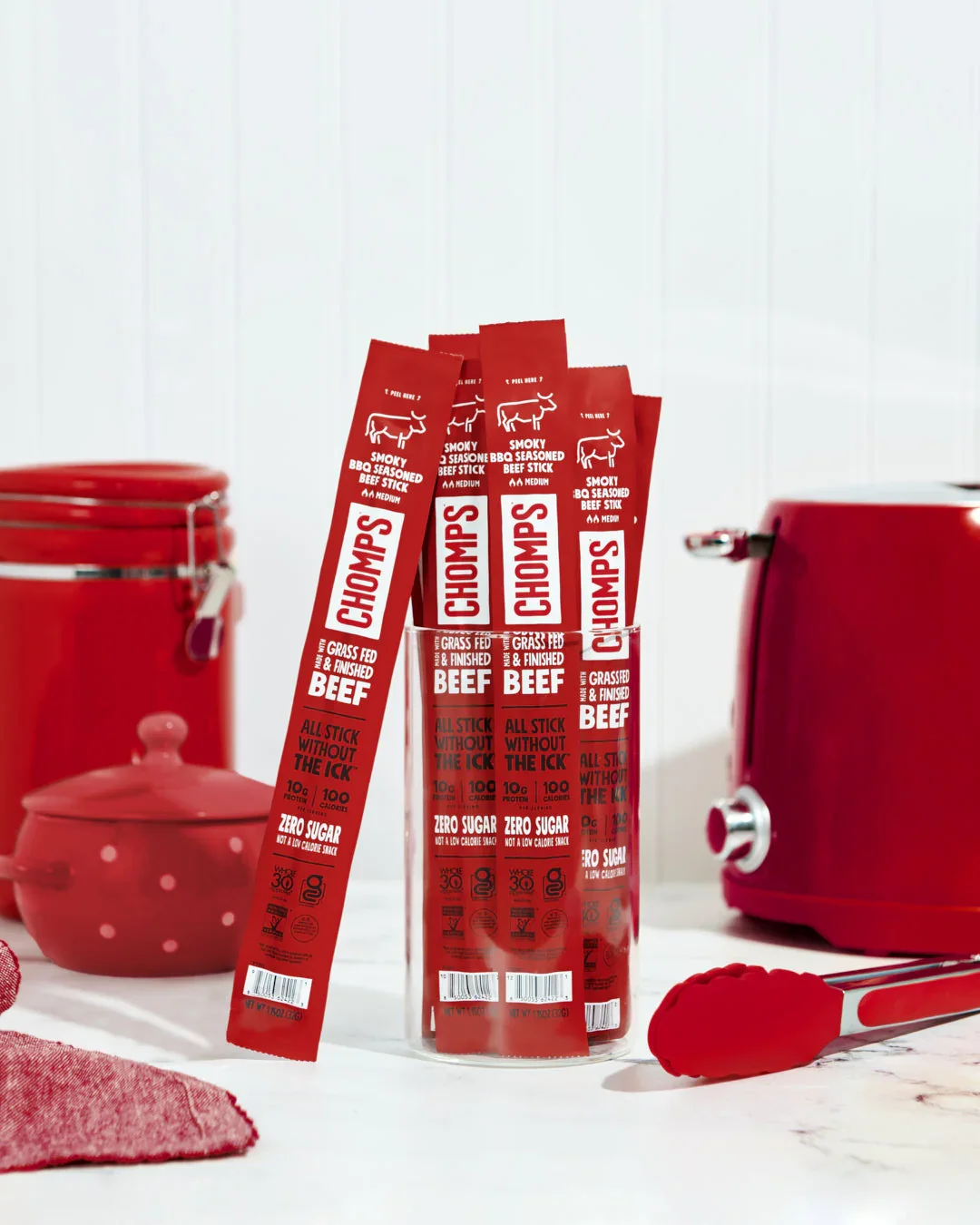 Red packages of Chomps beef sticks in a clear container with red kitchen items and a white background.