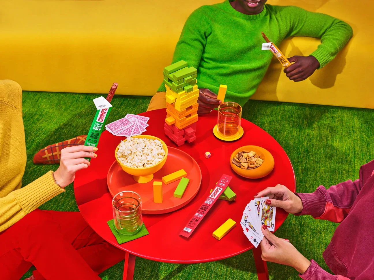 Three people playing a game of Jenga and cards at a red table with snacks and drinks.