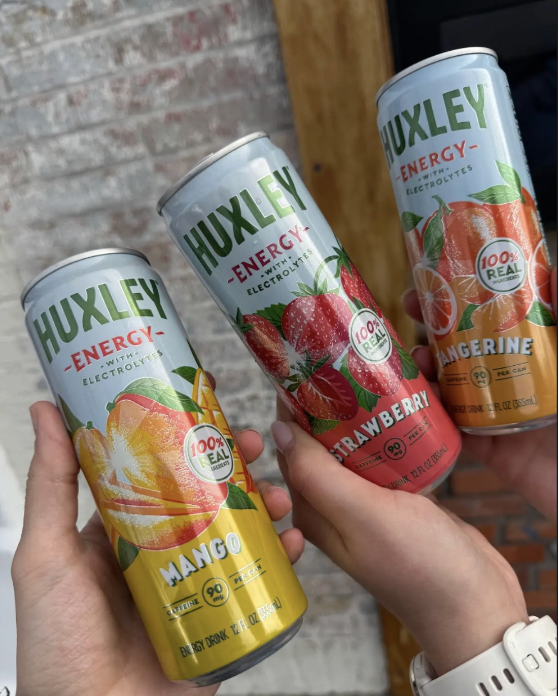 Three cans of Huxley Energy drinks with fruit flavors: mango, strawberry, and tangerine, held in hands.