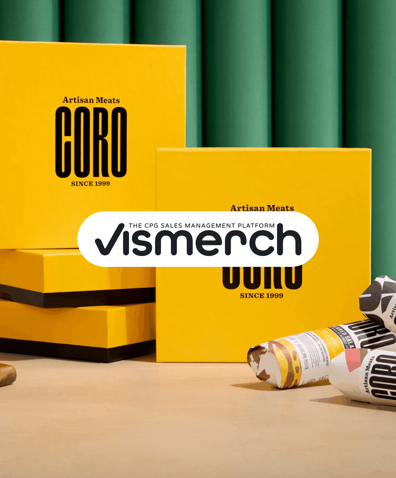 Stacked yellow boxes with black text reading "Artisan Meats Guro Since 1999" and a logo overlay for vismerch in black and white, with green backdrop and some rolled product on the table.