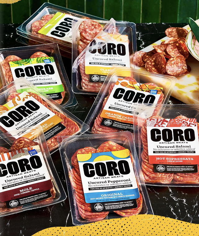 Multiple packages of CORO artisan meats including salami, pepperoni, and hot soppressata, arranged on a black surface. There is also a plate of sliced salami and a yellow patterned item partially visible at the bottom.