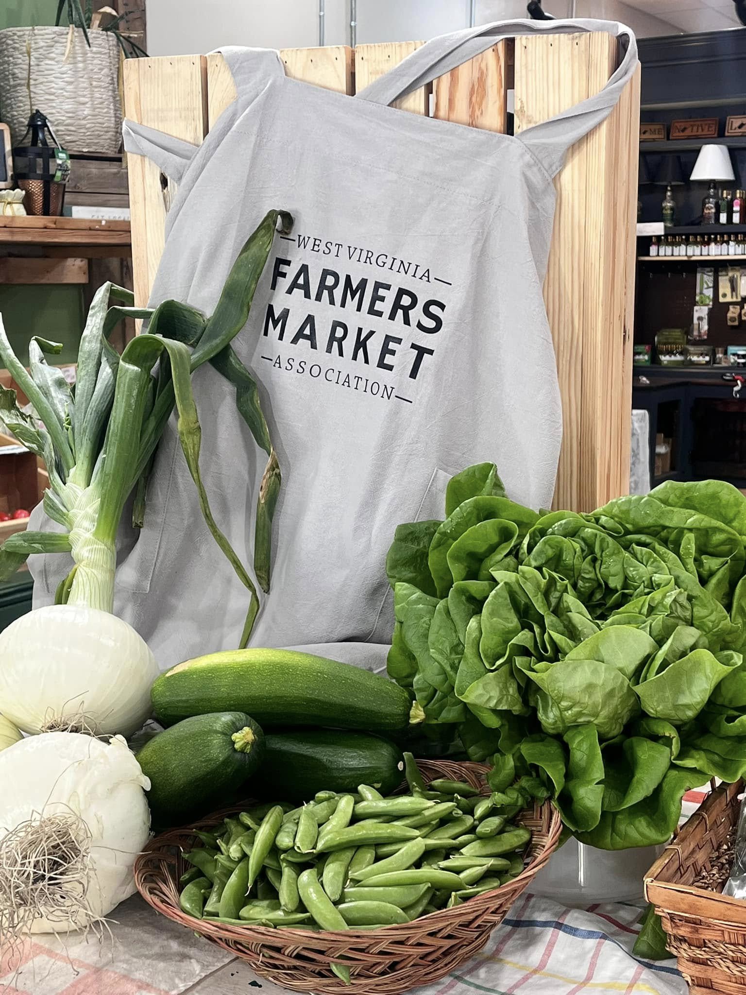 A display of fresh vegetables including onions, zucchini, peas, and lettuce, with a West Virginia Farmers Market Association apron in the background.