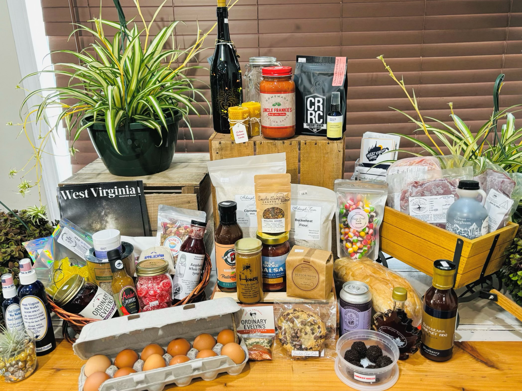 Assortment of West Virginia local products and plants on a wooden table, including artisanal sauces, baked goods, jarred items, eggs, and a West Virginia magazine.