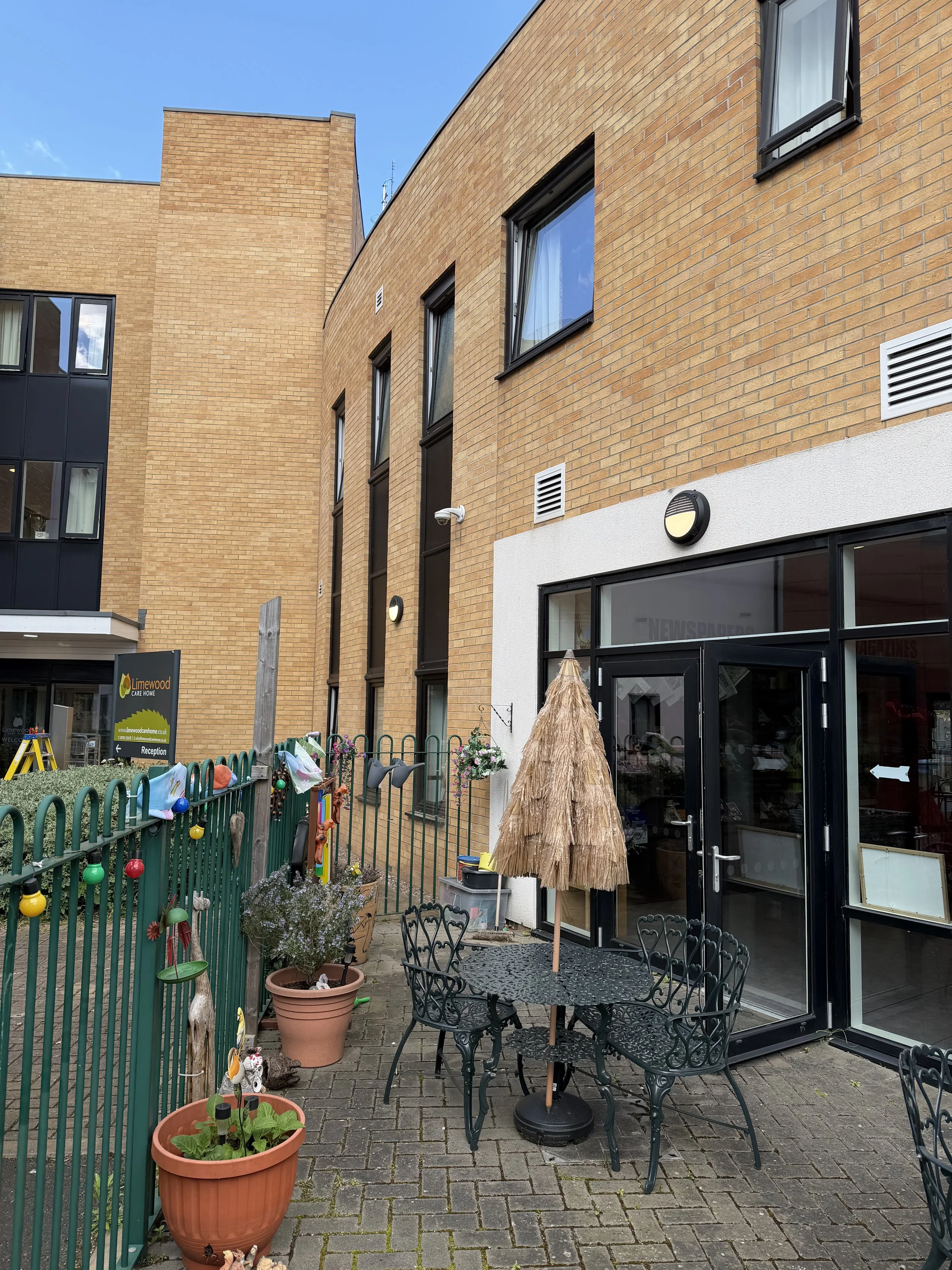 A small outdoor patio area with a black metal table and four matching chairs, an umbrella with a straw-like cover, potted plants, a green metal fence with colorful hanging decorations, and the exterior of a brick building with large windows and a gla